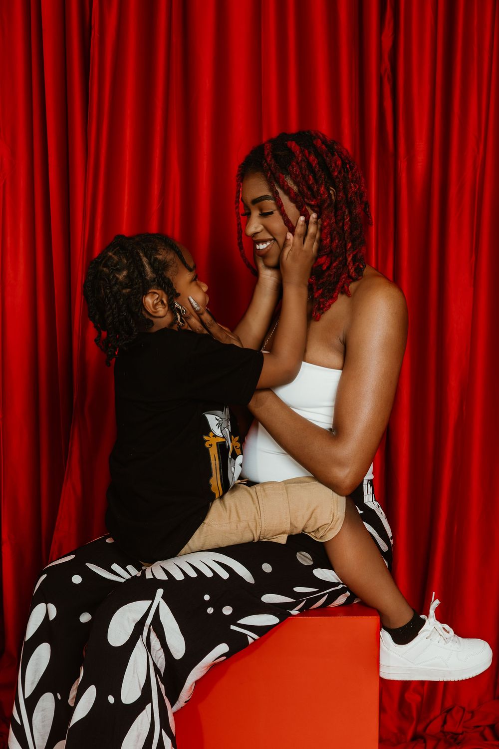 A joyful Black mother and child share a tender moment on a red cube against a vibrant red curtain backdrop.