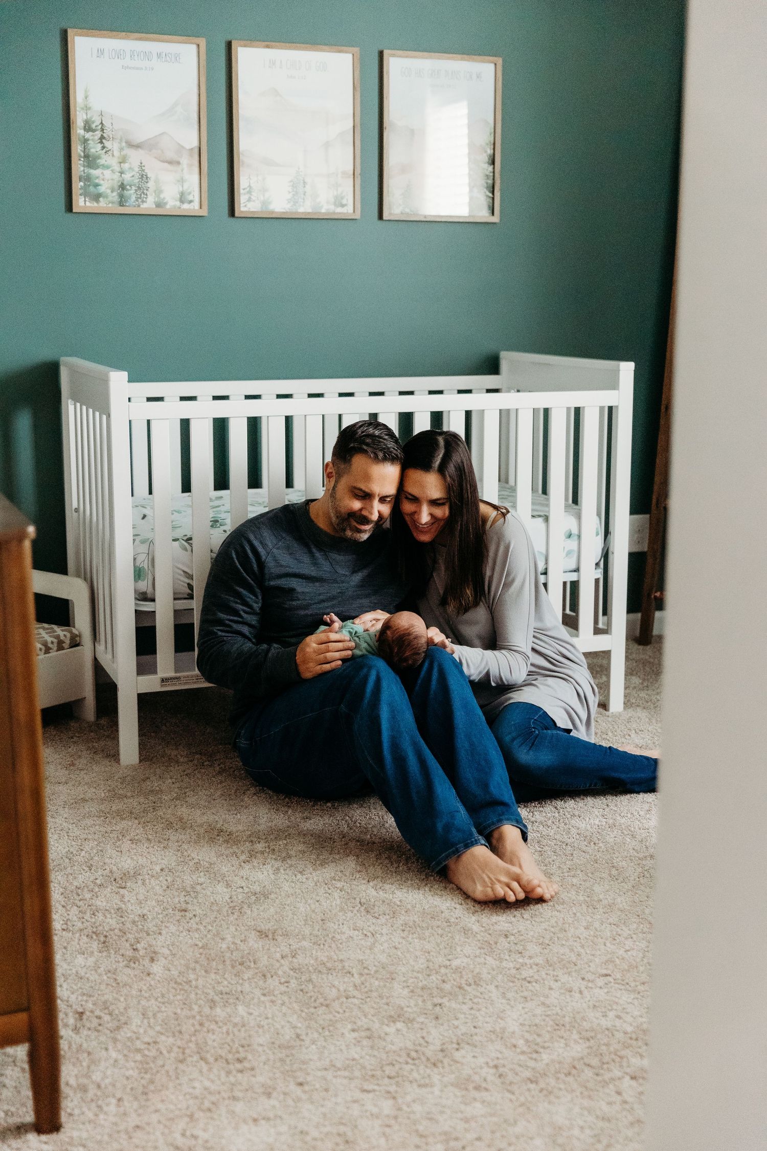 Couple sitting on floor looking at phone in nursery with white crib and green walls.