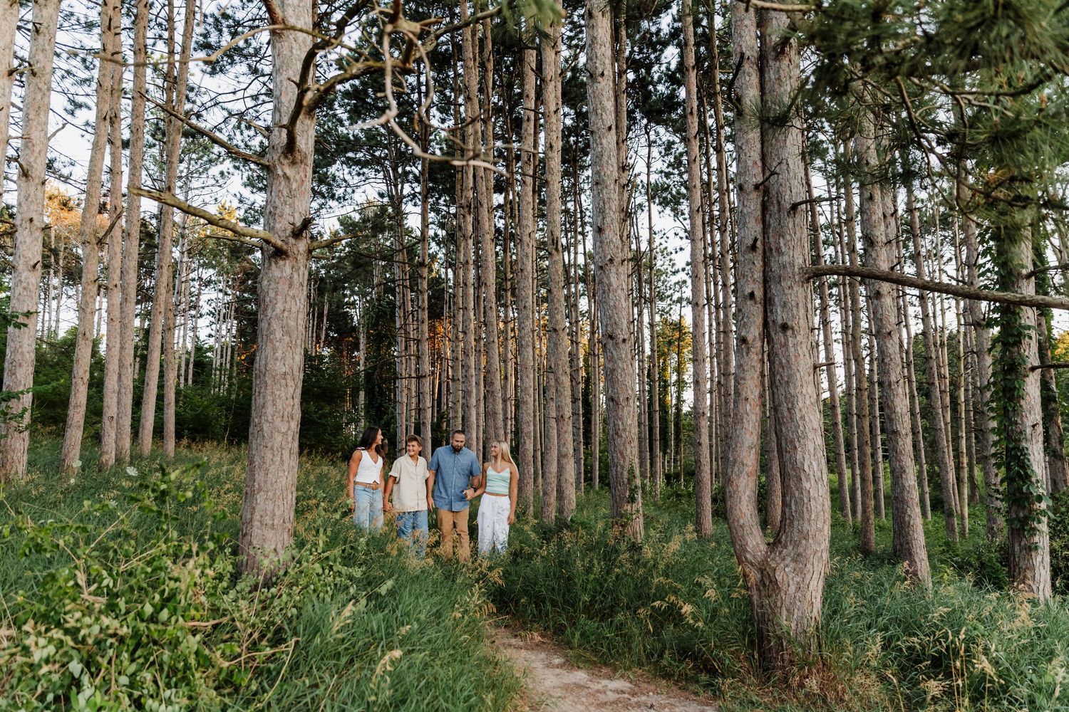 Family of four walking on a forest path surrounded by tall pine trees and lush green foliage.