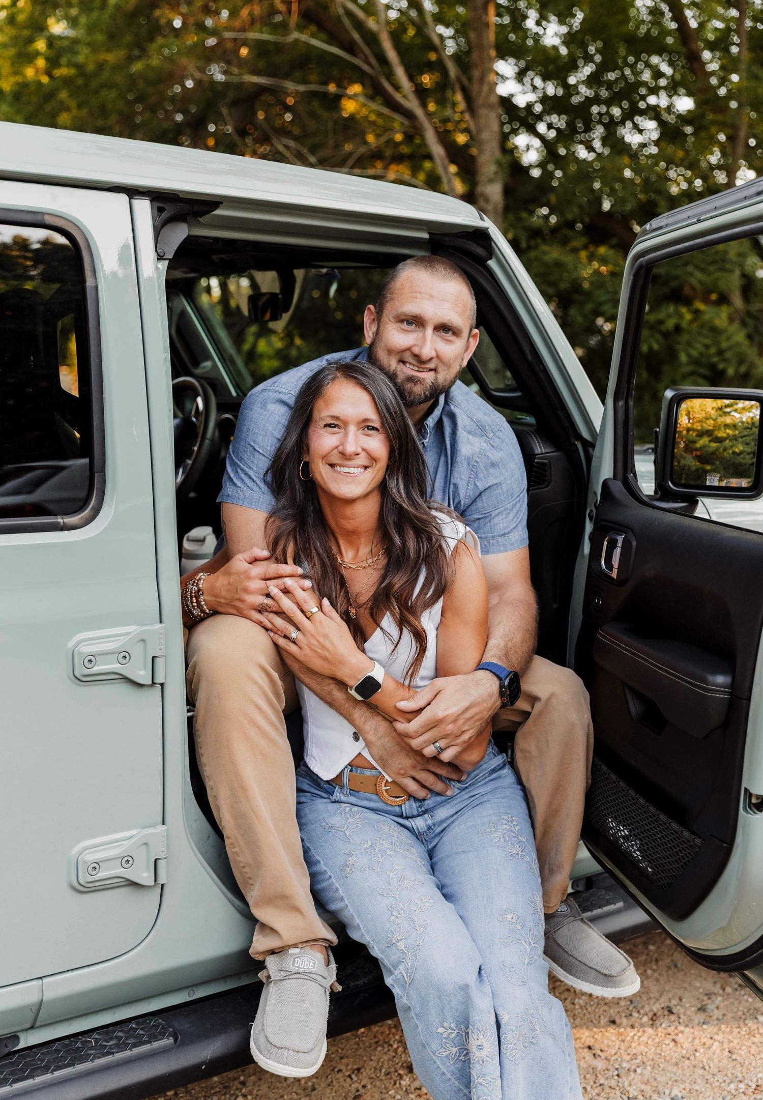 A smiling couple poses together in the open door of a mint green Jeep Wrangler, embracing warmly outdoors.