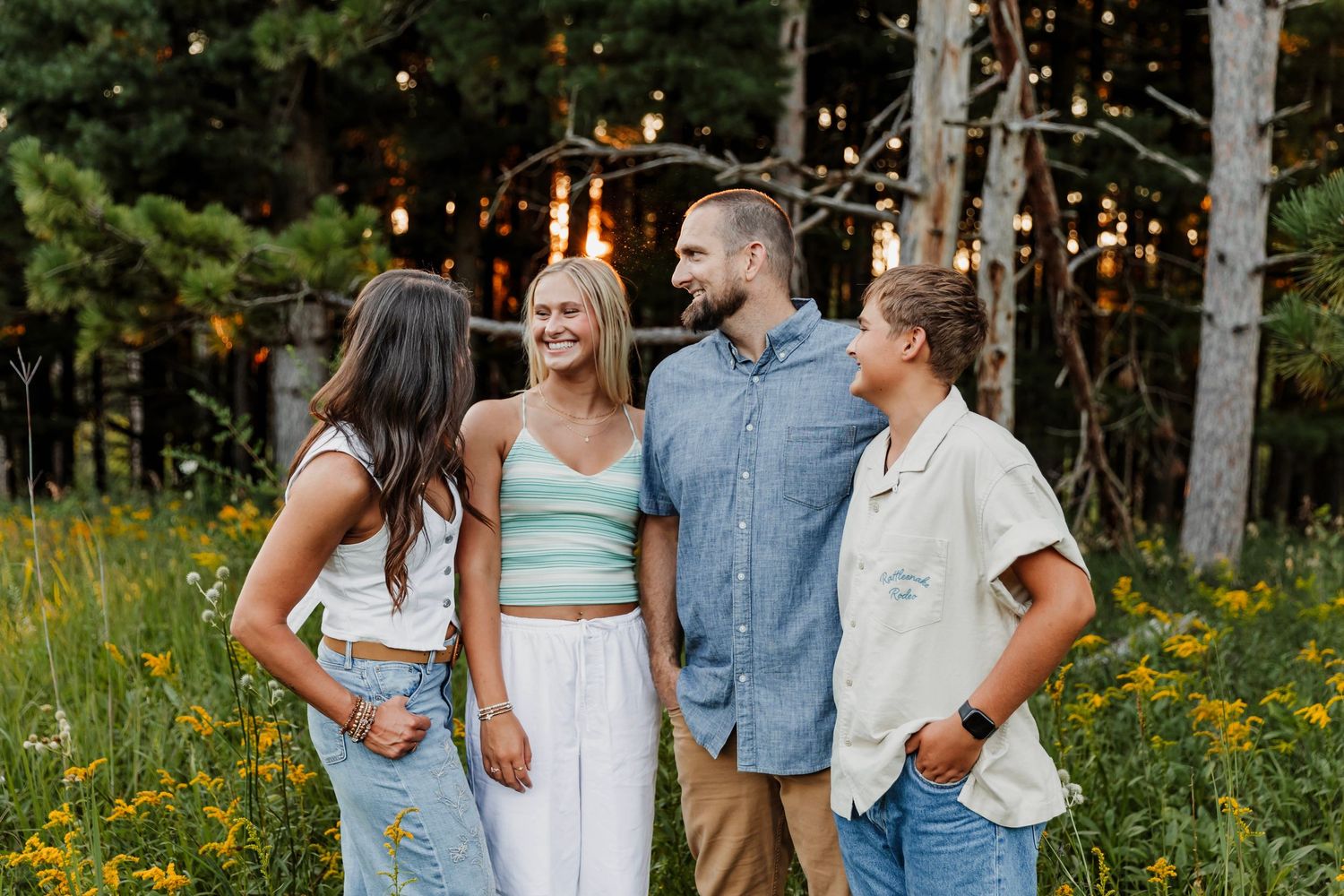 Four friends laughing together outdoors among wildflowers and string lights at golden hour.
