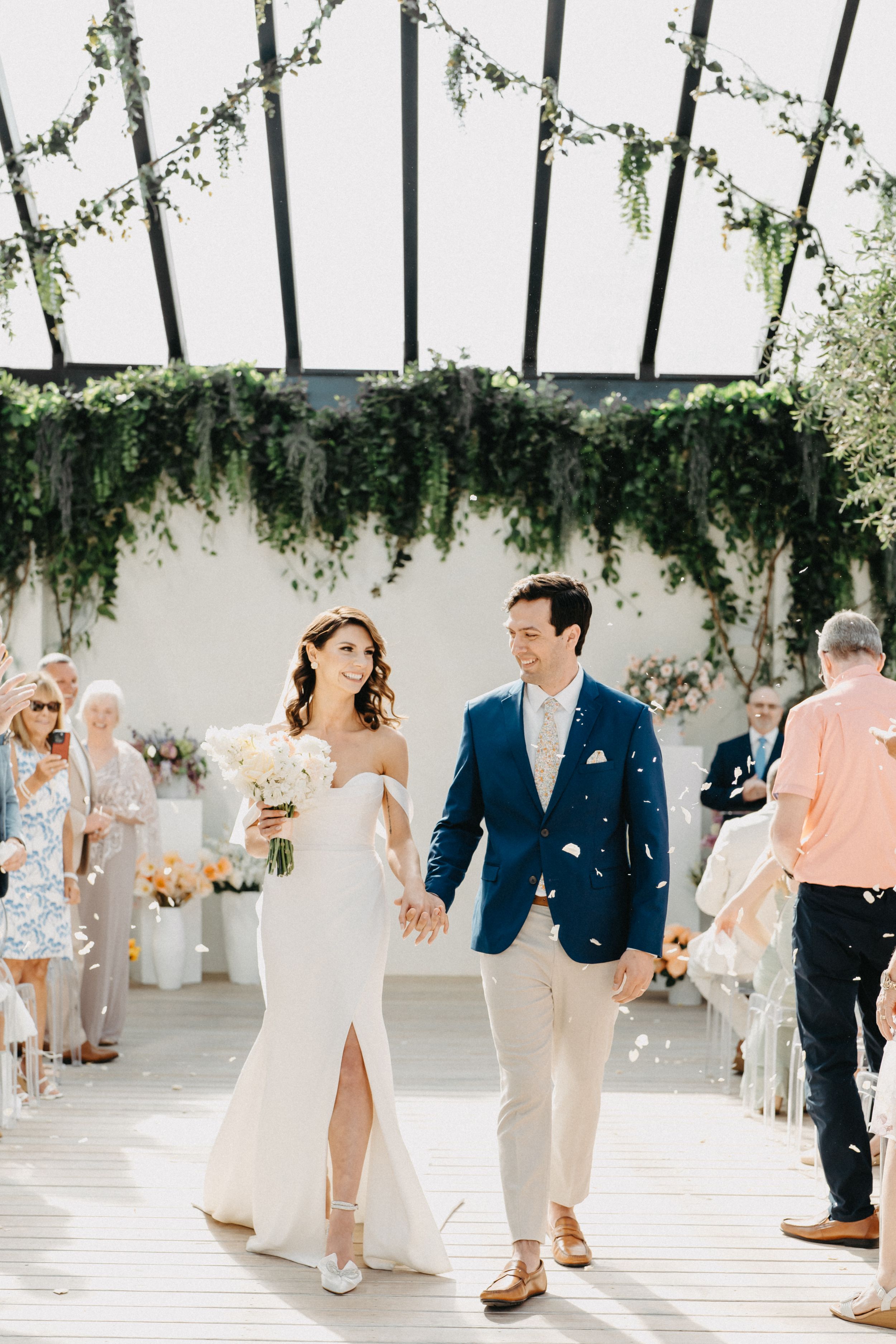 Bride and groom walk down the aisle hand in hand, smiling, in a sunlit greenhouse venue adorned with lush greenery.