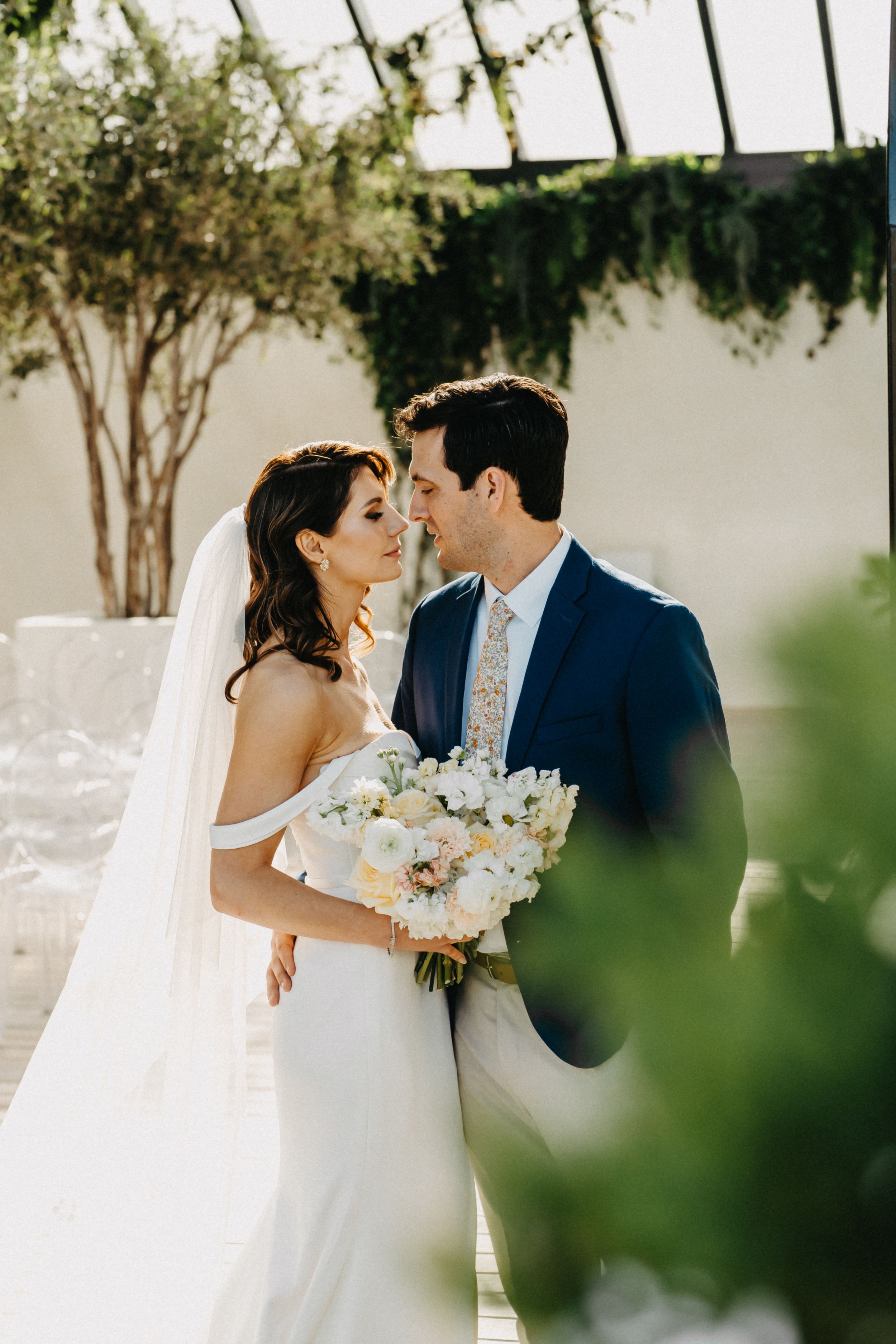 Bride in off-shoulder gown holding white bouquet shares romantic moment with groom in navy suit outdoors.