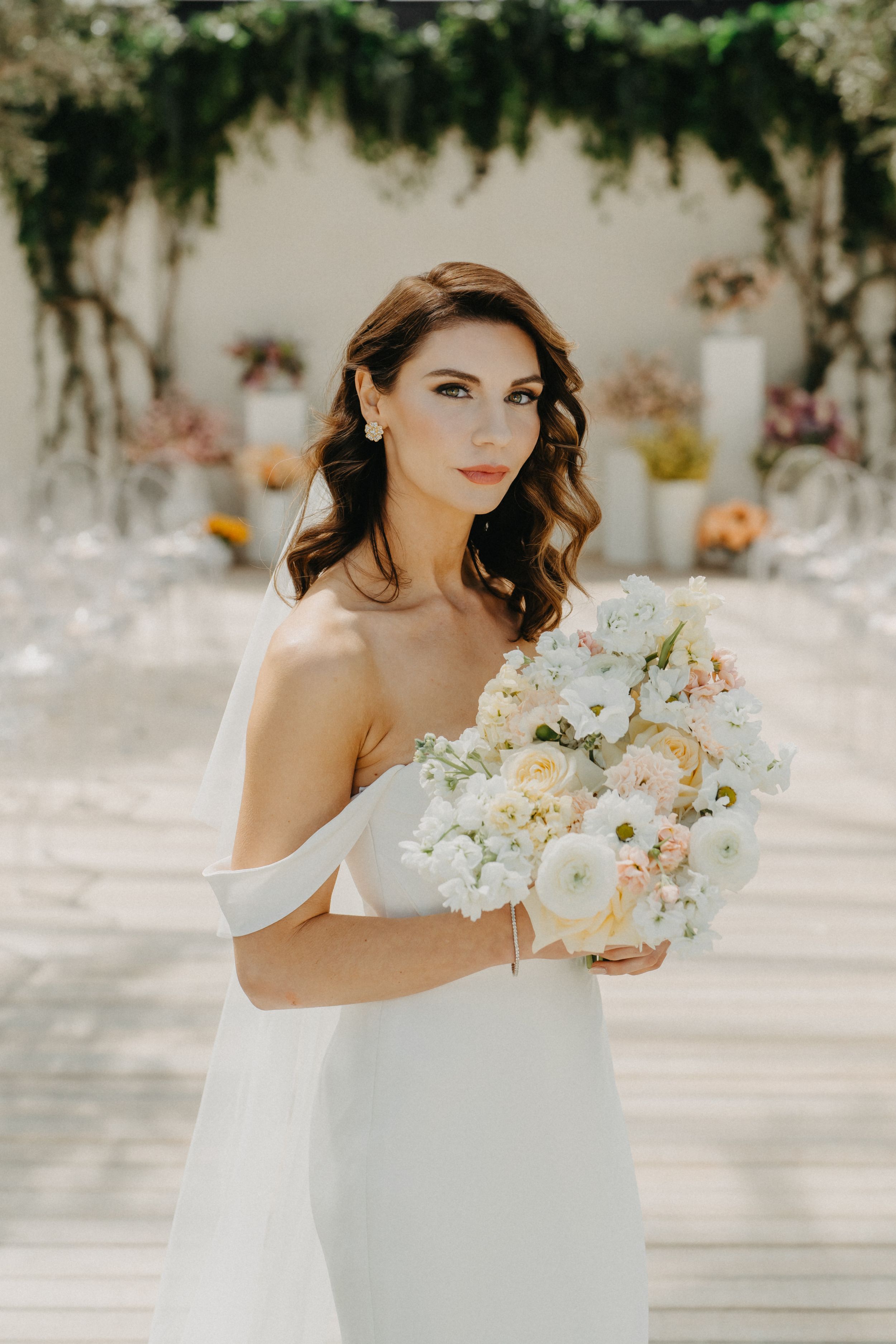 Elegant bride in off-shoulder white gown holding a lush bouquet of white and blush flowers at an outdoor wedding venue.