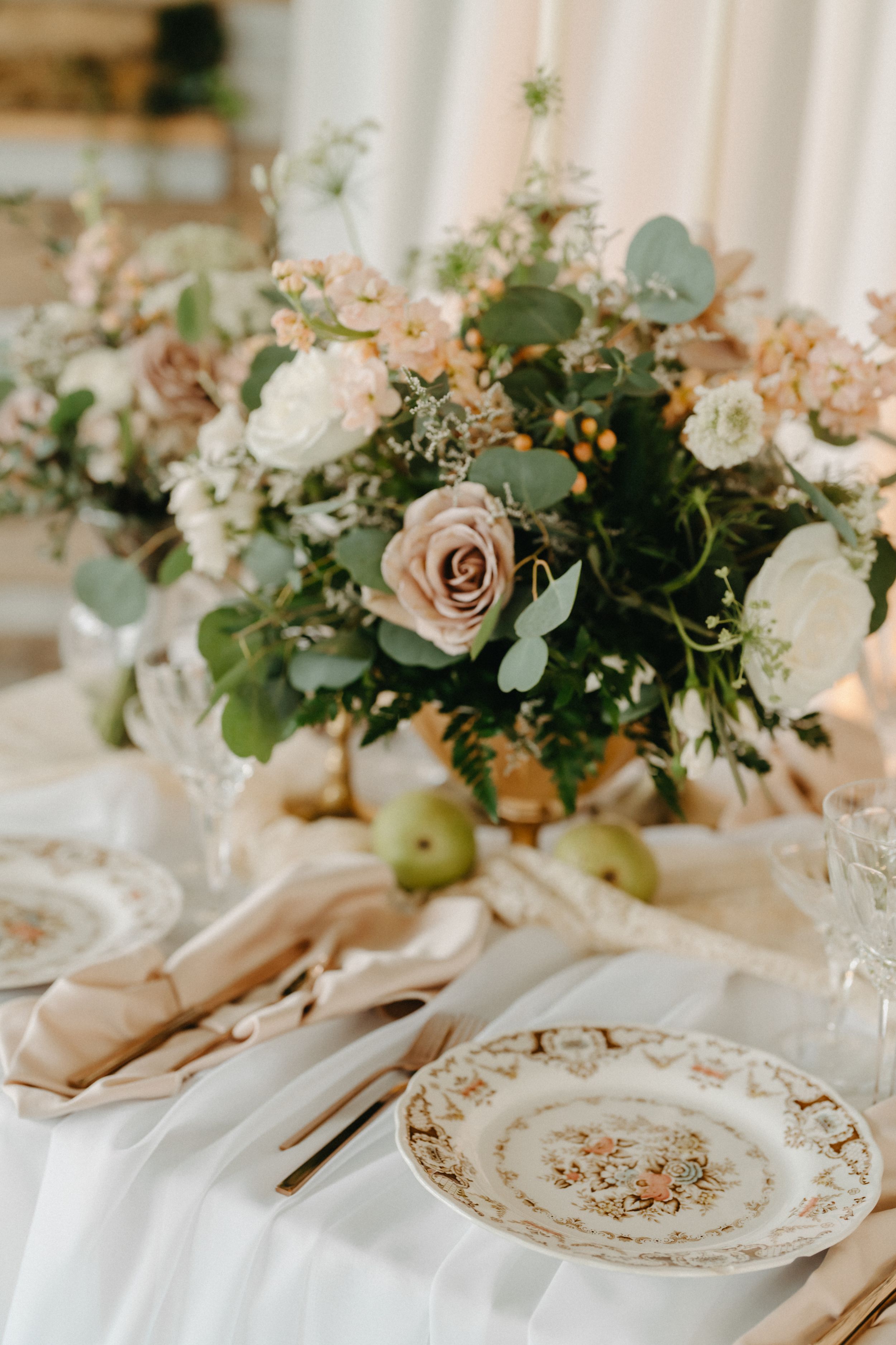 Elegant wedding table setting with vintage floral plates, gold cutlery, linen napkins, and a lush centerpiece of roses and eucalyptus.