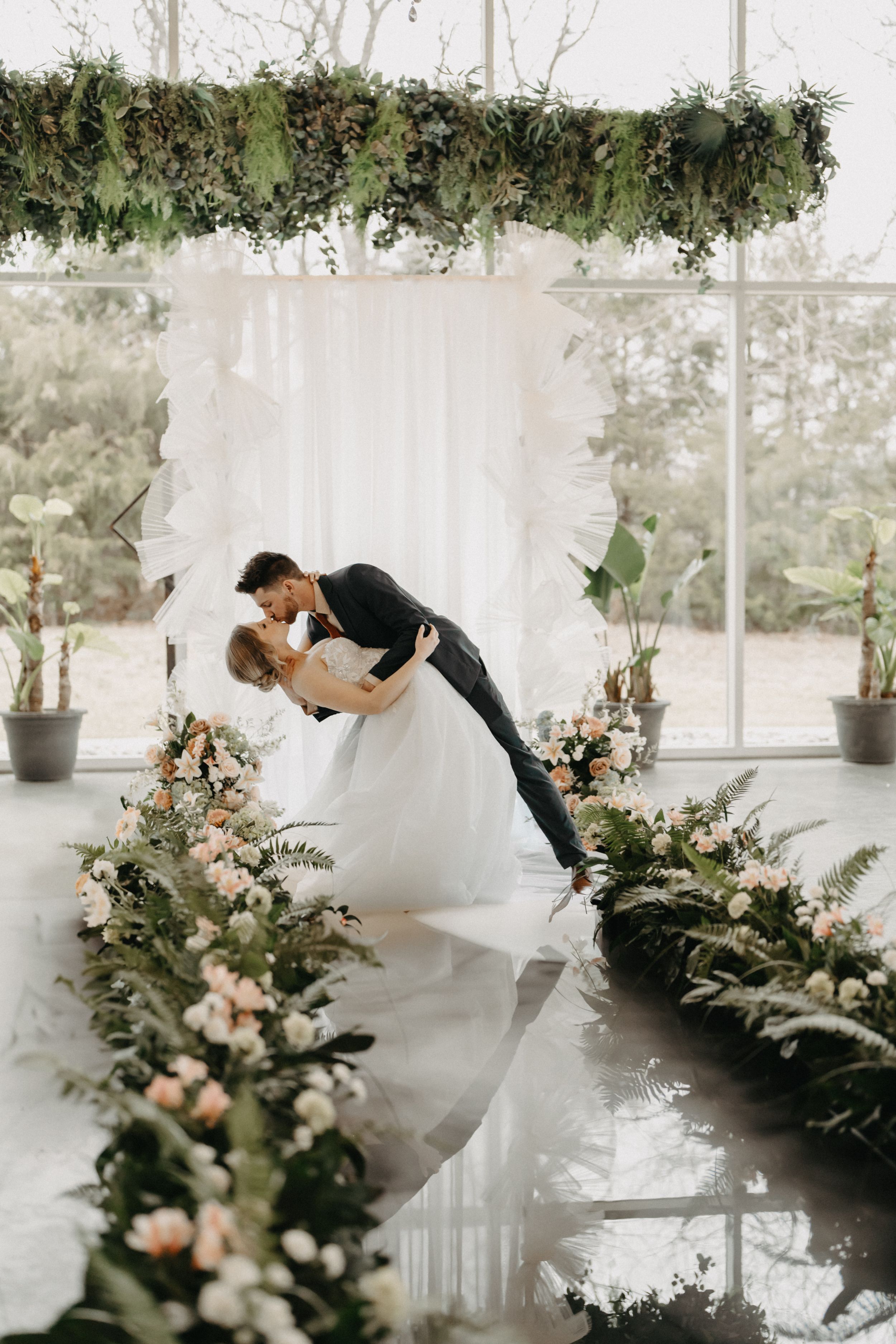 Bride and groom share a romantic dip kiss at a floral wedding altar with lush greenery and pink blooms.