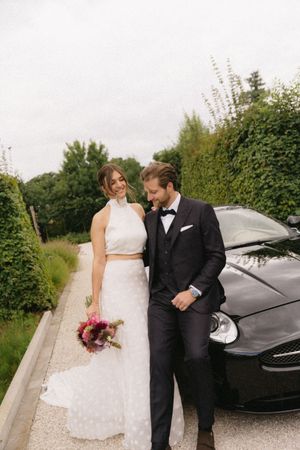 A wedding couple poses next to a sleek black car outdoors.