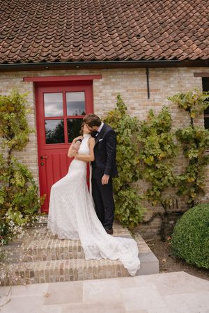 A wedding couple shares an intimate moment by a rustic brick building with a red door.