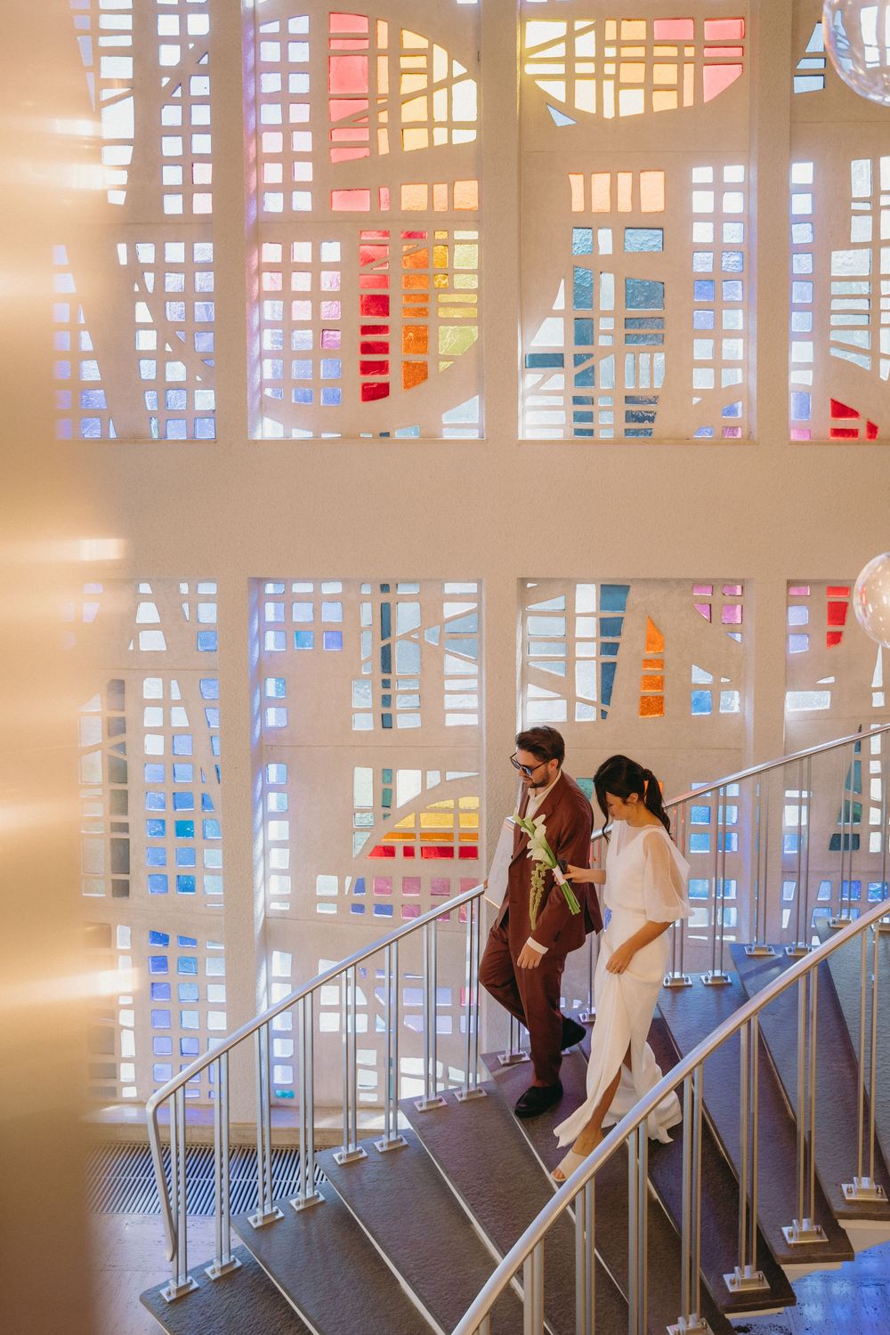 Couple ascending modern stairs with colorful stained glass windows creating a dramatic backdrop.