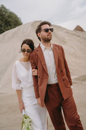 A couple in white dress and brown suit poses together against desert hills.