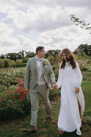 A couple in wedding attire strolls through a blooming rose garden on a cloudy day.