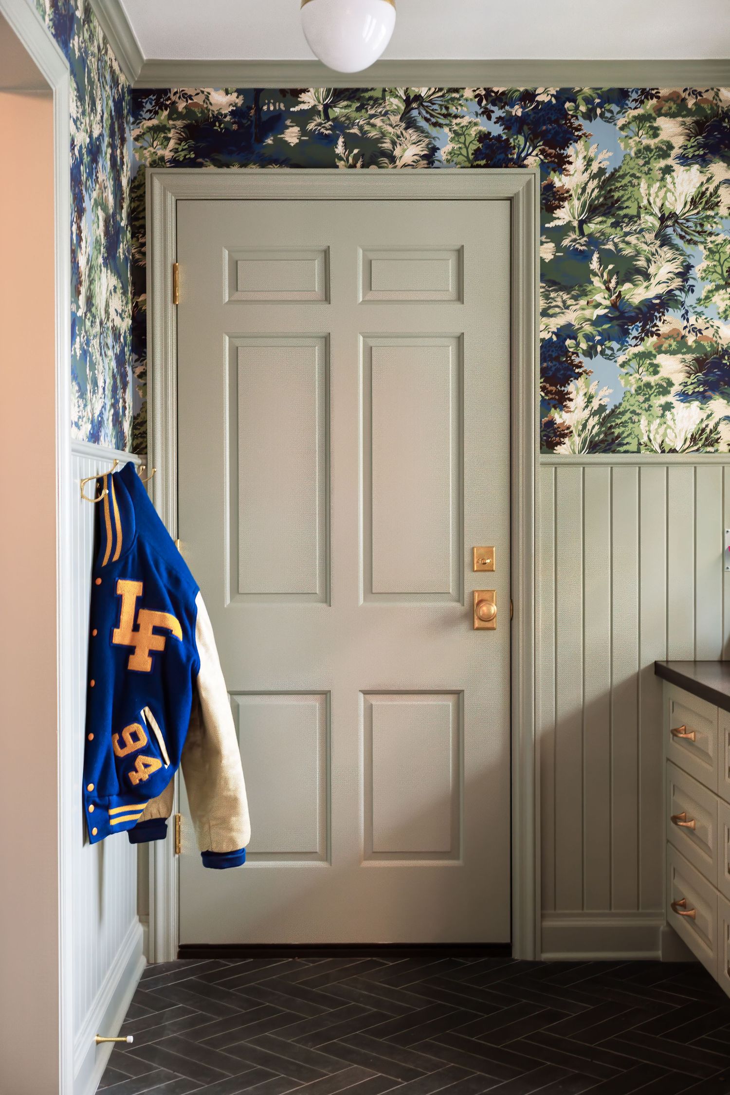 A gray door with floral wallpaper, wainscoting, and a varsity letterman jacket hanging on a hook, featuring dark herringbone flooring.