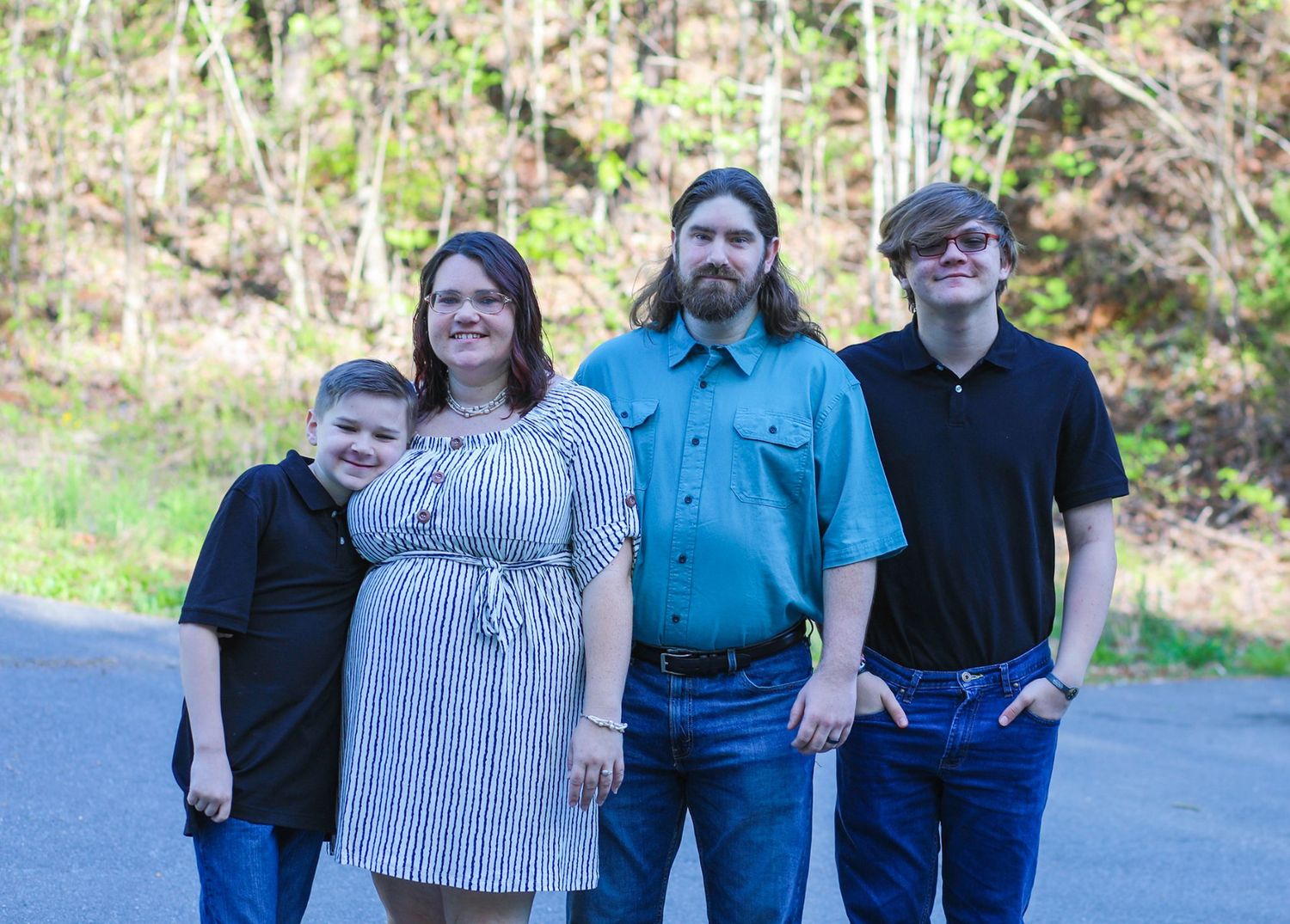 Family of four posing outdoors on a road surrounded by lush green trees in a casual portrait photo.