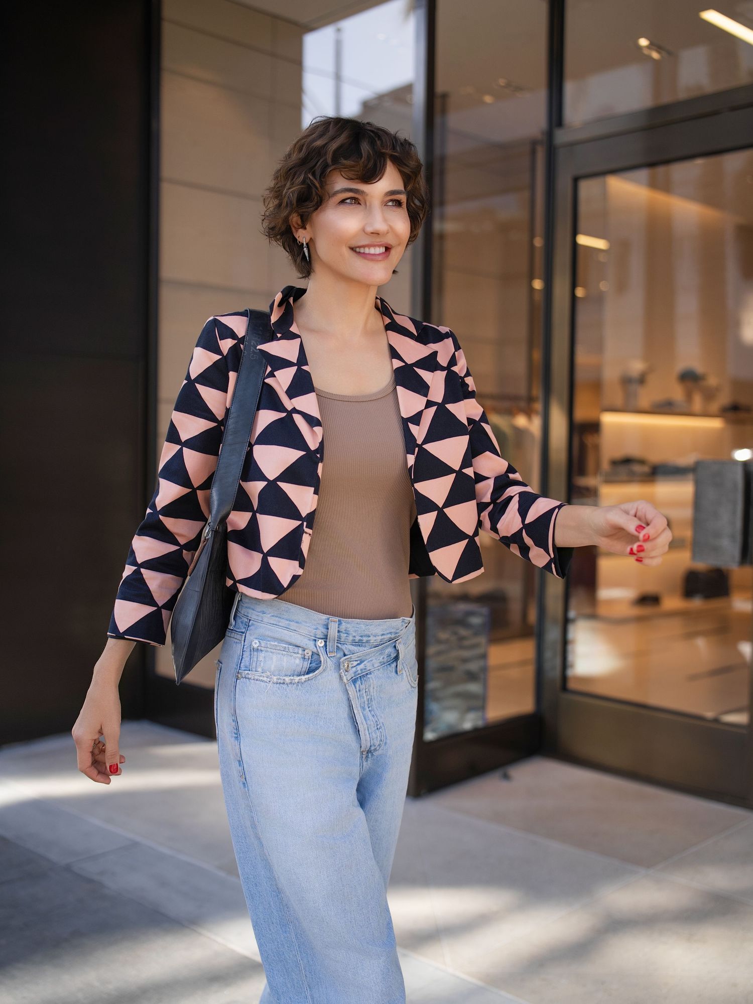 Woman in pink geometric blazer, tan top, and light jeans carries black bag outside a modern storefront.