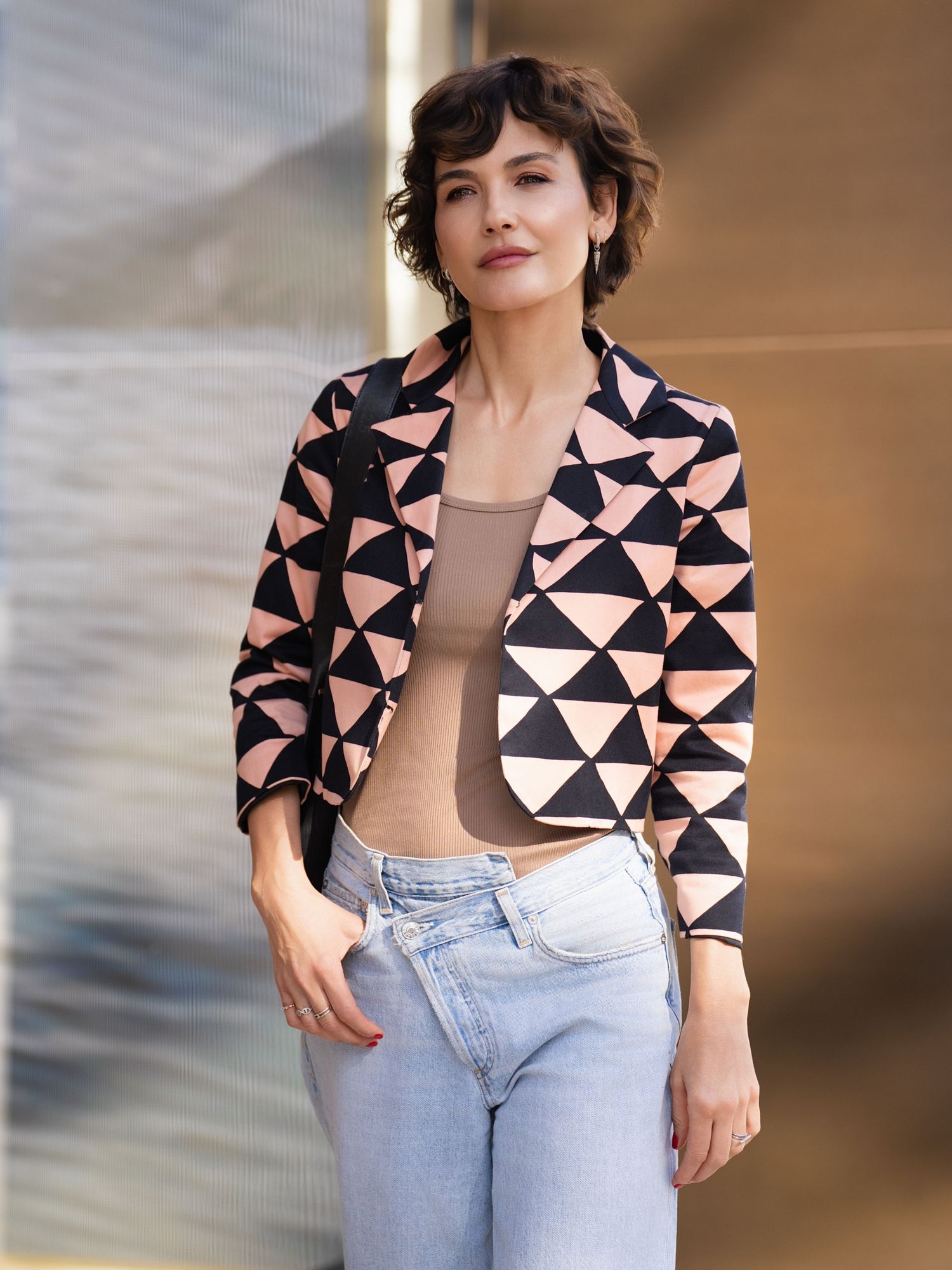 Woman wearing a geometric print blazer in pink and black over a beige top and light wash jeans.