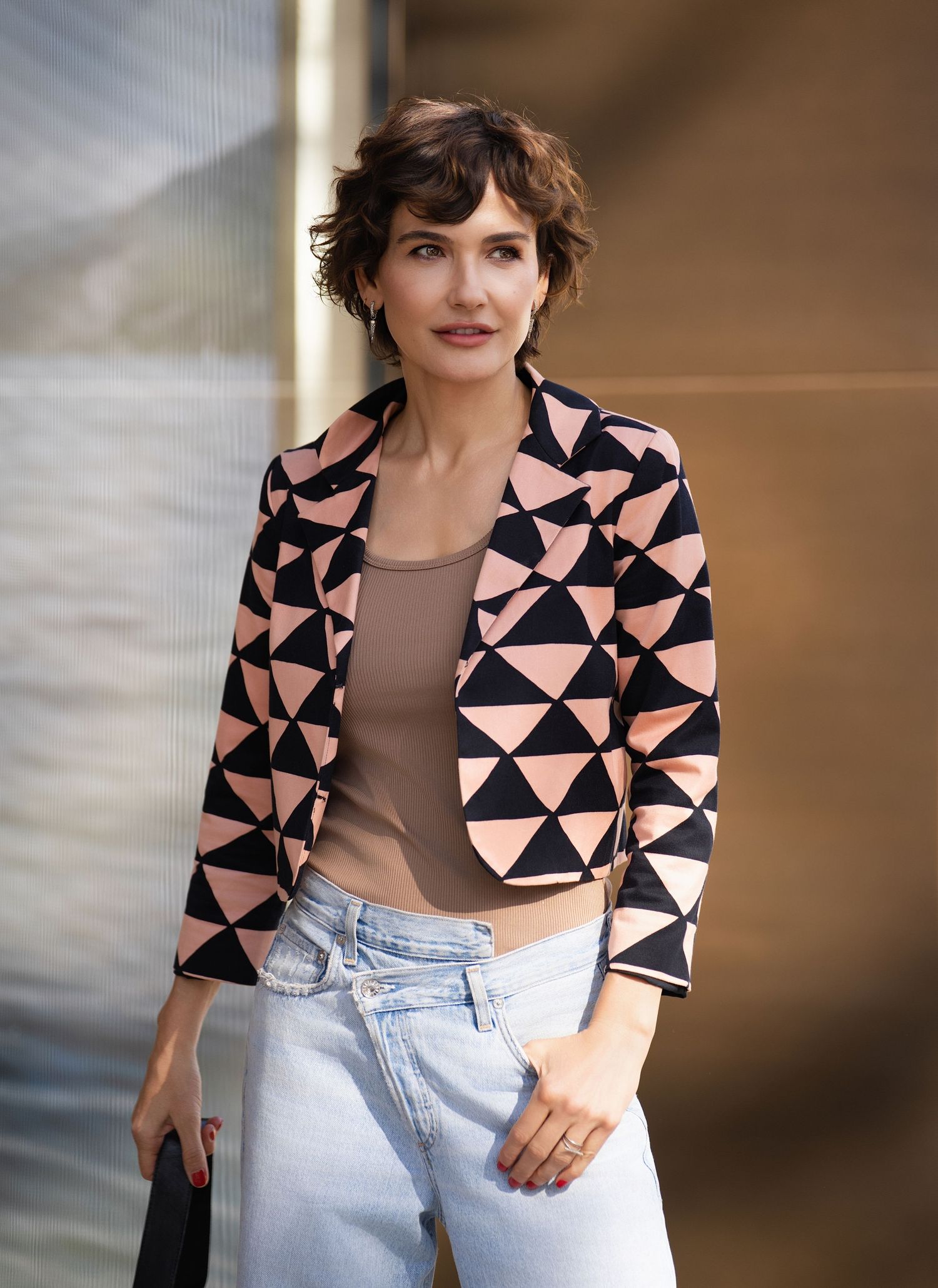 Woman in geometric black and pink blazer with light jeans, short curly hair, posing outdoors.