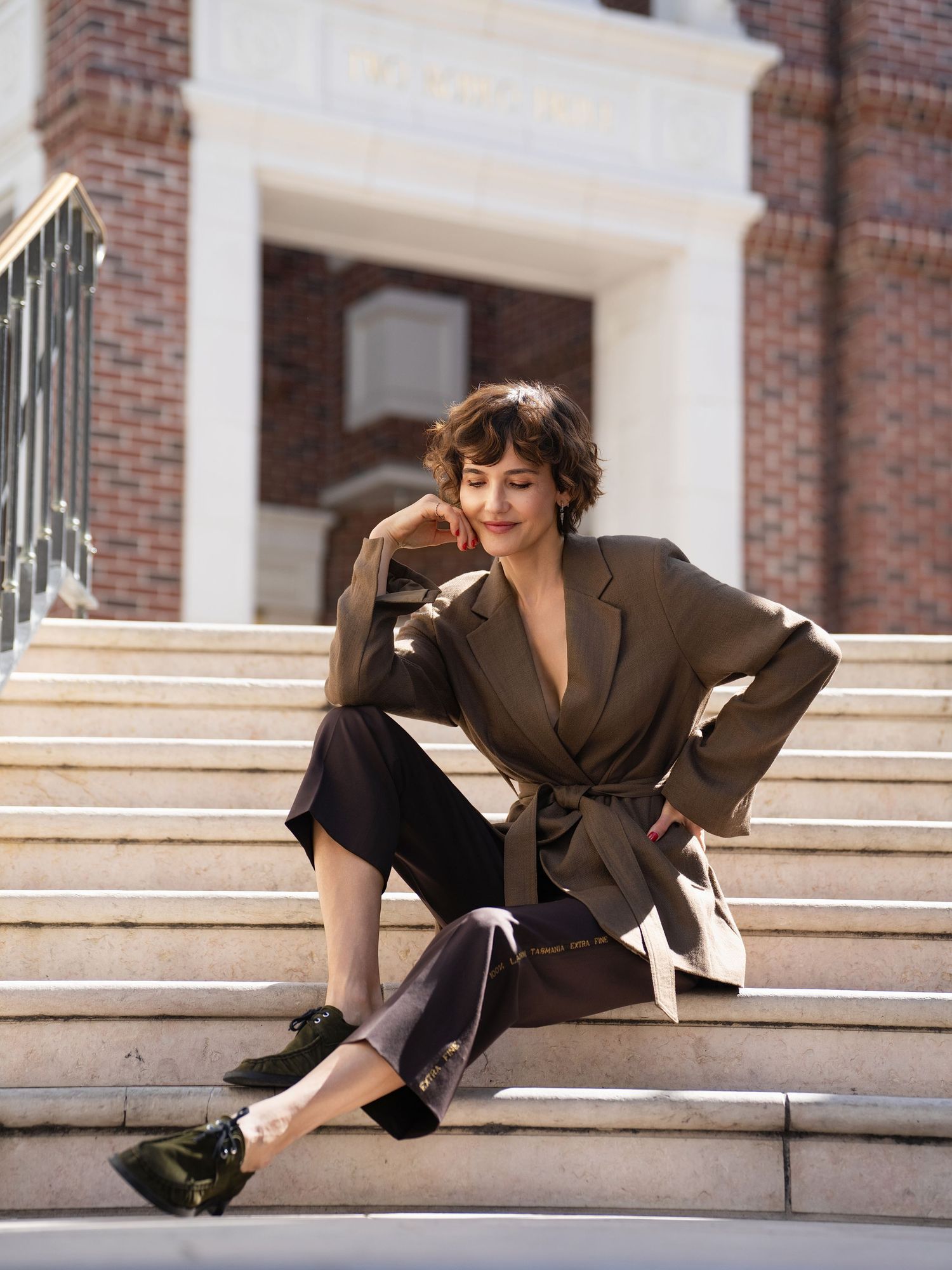 Woman in olive belted blazer and dark trousers sits on marble steps outside a brick building.