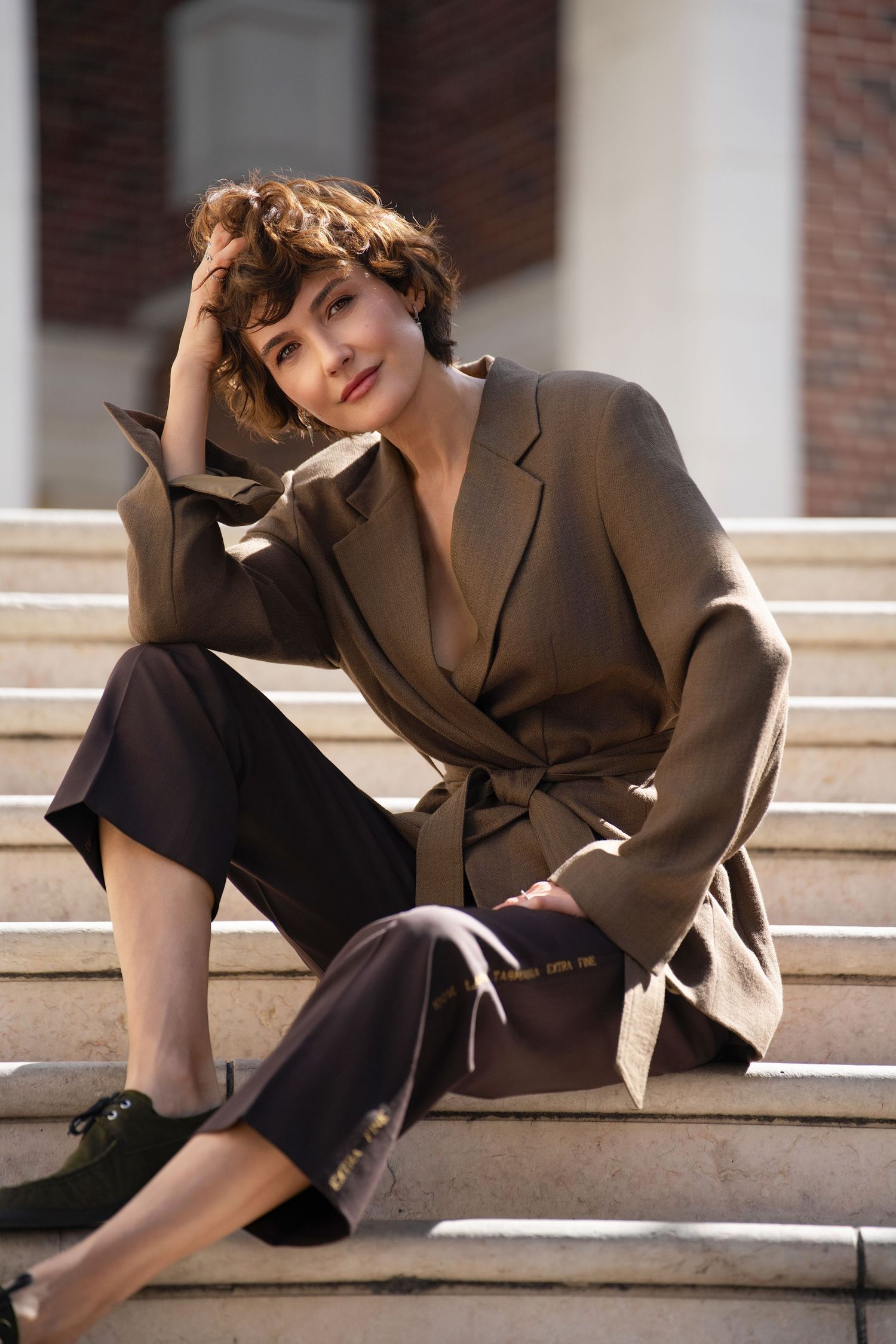 Woman in olive wrap blazer and brown trousers sitting on stone steps outdoors.