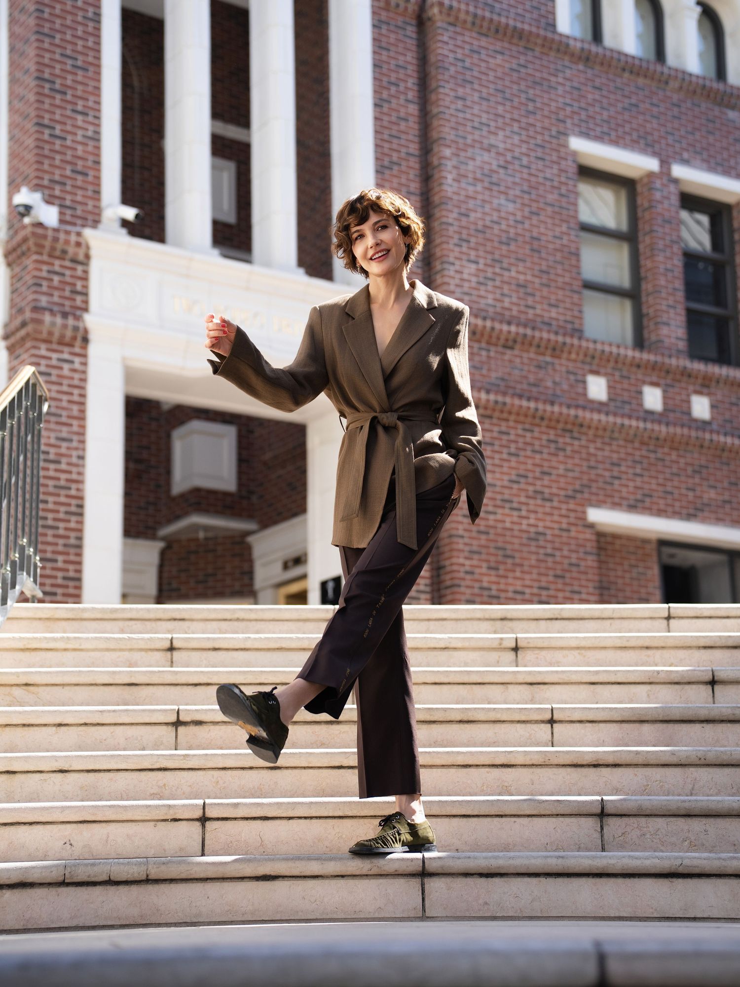 Woman in olive belted blazer and dark trousers poses on sunlit brick building steps.