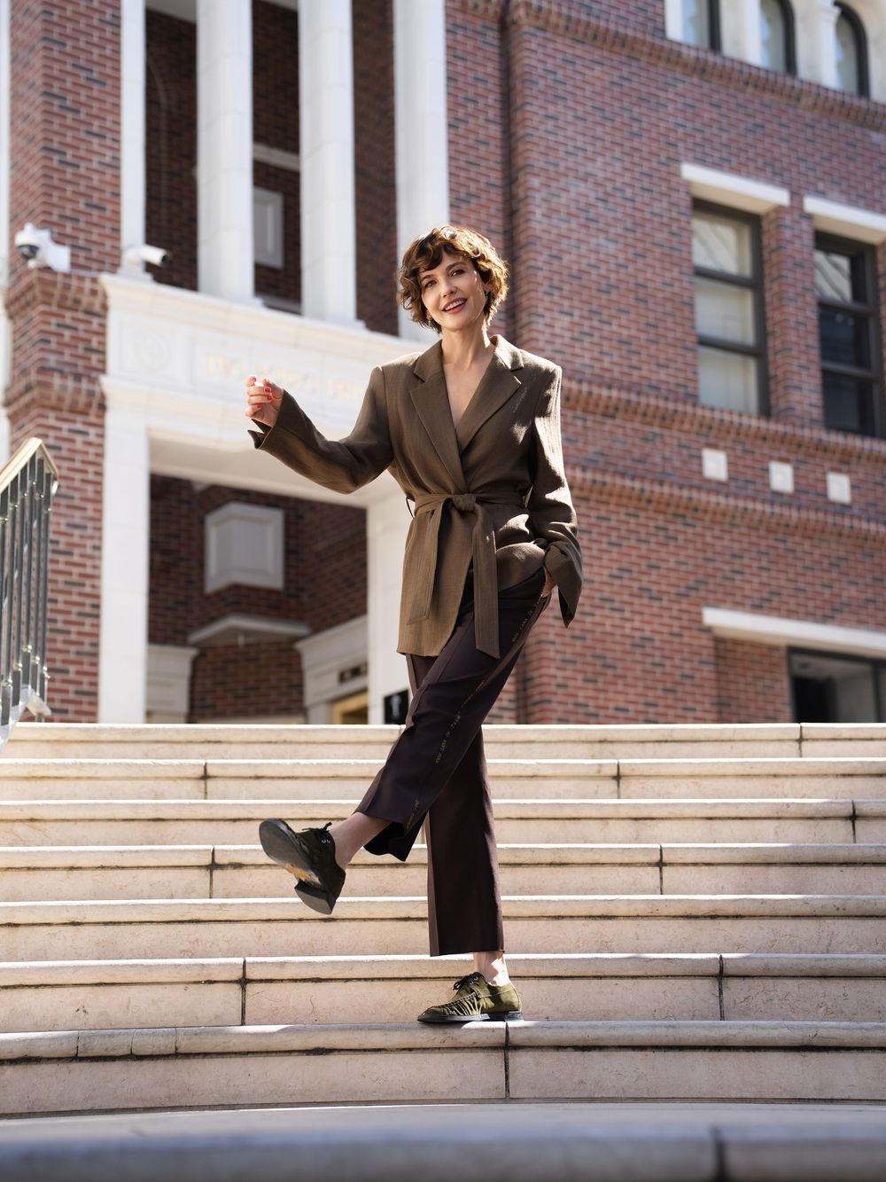 Woman in olive belted blazer and dark trousers poses on sunlit brick building steps.
