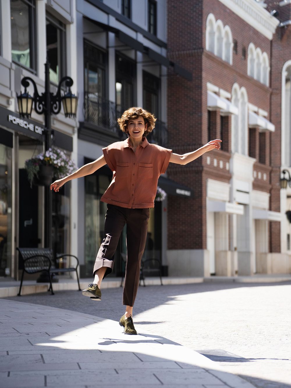 Person with curly hair joyfully dancing on a sunny urban street, wearing a rust shirt, dark pants, and green shoes.