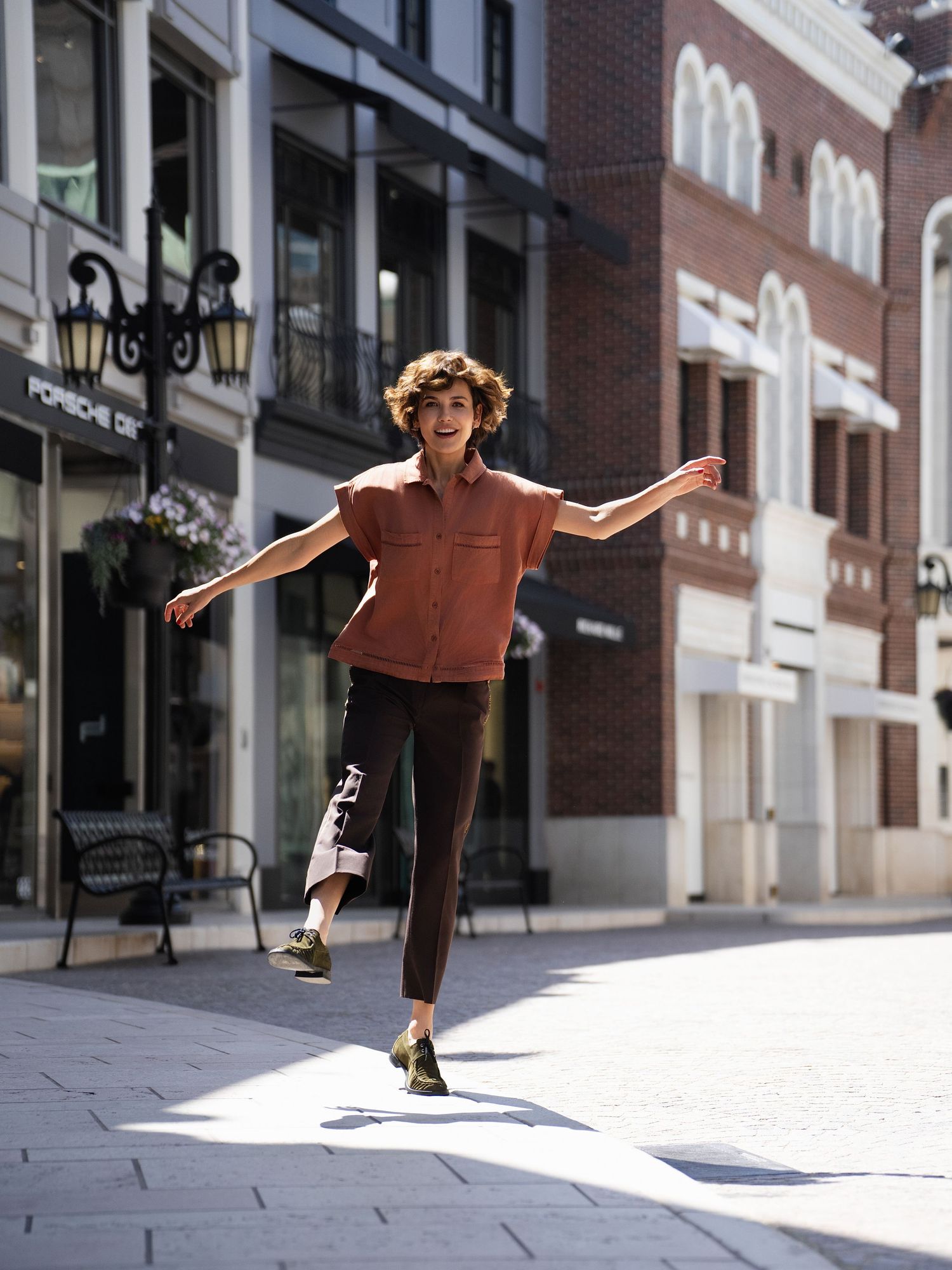 Person with curly hair joyfully dancing on a sunny urban street, wearing a rust shirt, dark pants, and green shoes.