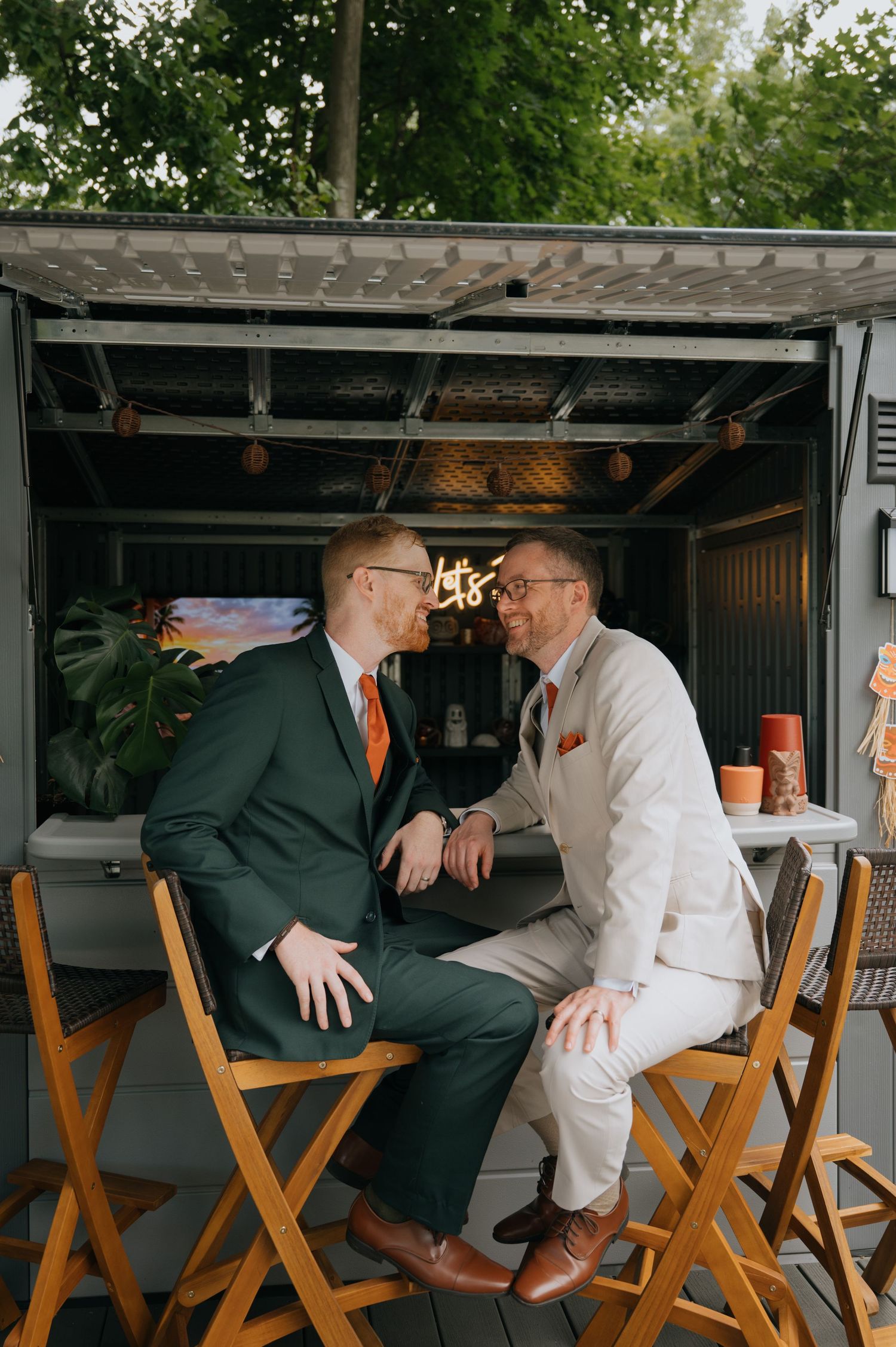 Two grooms in green and white suits share a romantic moment at an outdoor bar during their wedding reception.