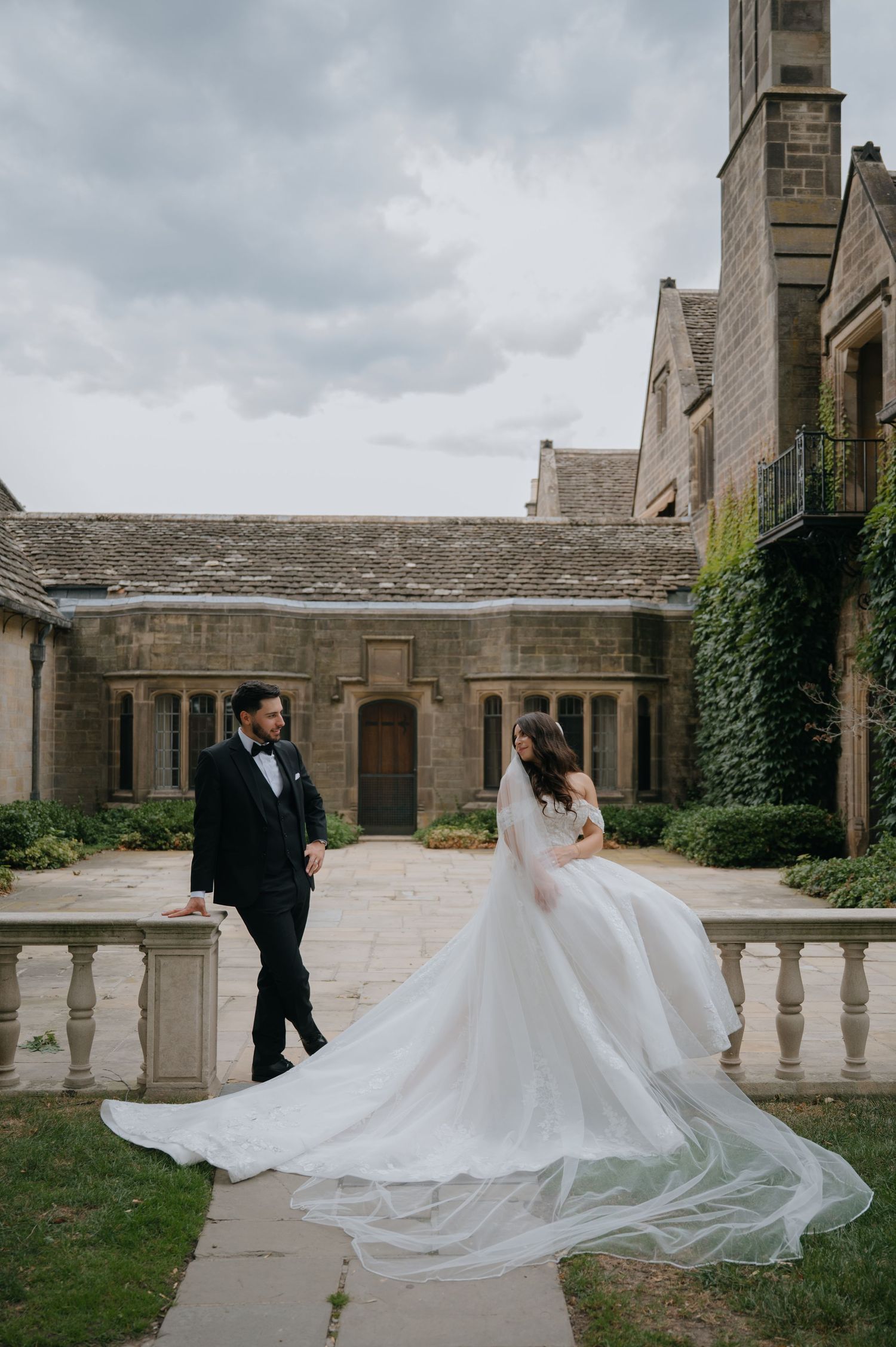 Bride in flowing white gown and groom in tuxedo walk through elegant stone courtyard of historic venue.