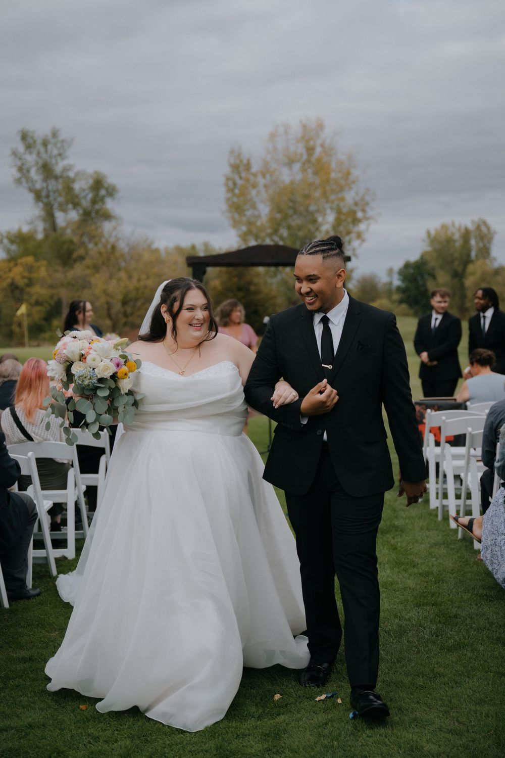 A wedding ceremony processional on a green lawn with white chairs and an overcast sky in the background.