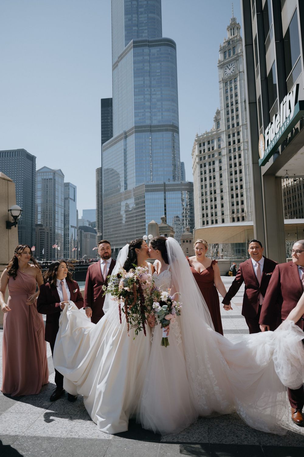 Bride and groom kiss on Chicago street with wedding party in mauve attire, Trump Tower skyline backdrop.