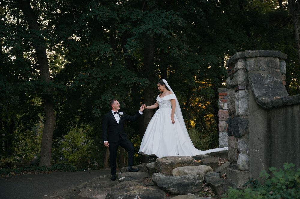A couple in formal wedding attire poses for photos in a scenic outdoor garden setting with stone architecture.