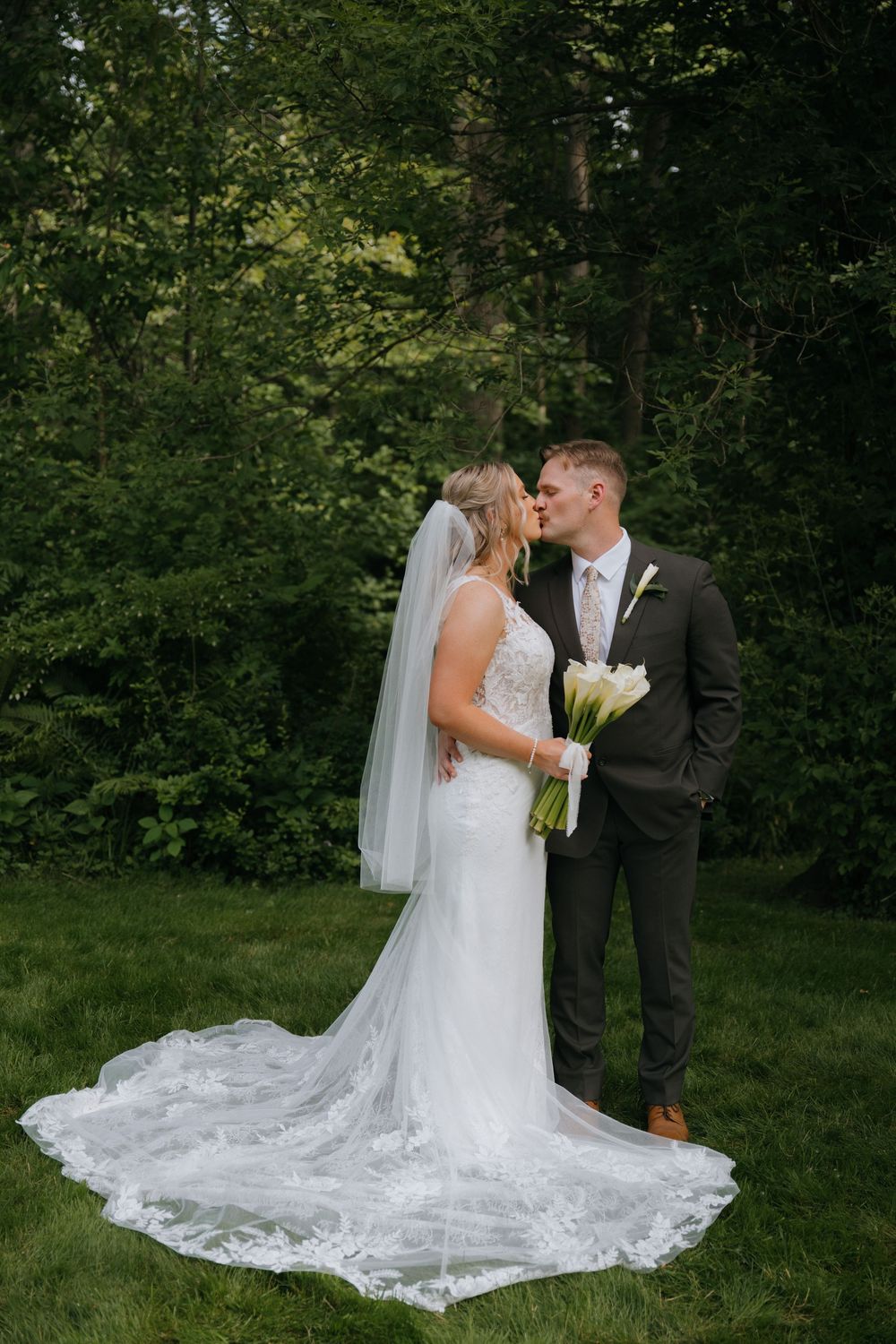 A bride and groom in formal wedding attire share intimate poses in a wooded outdoor setting.