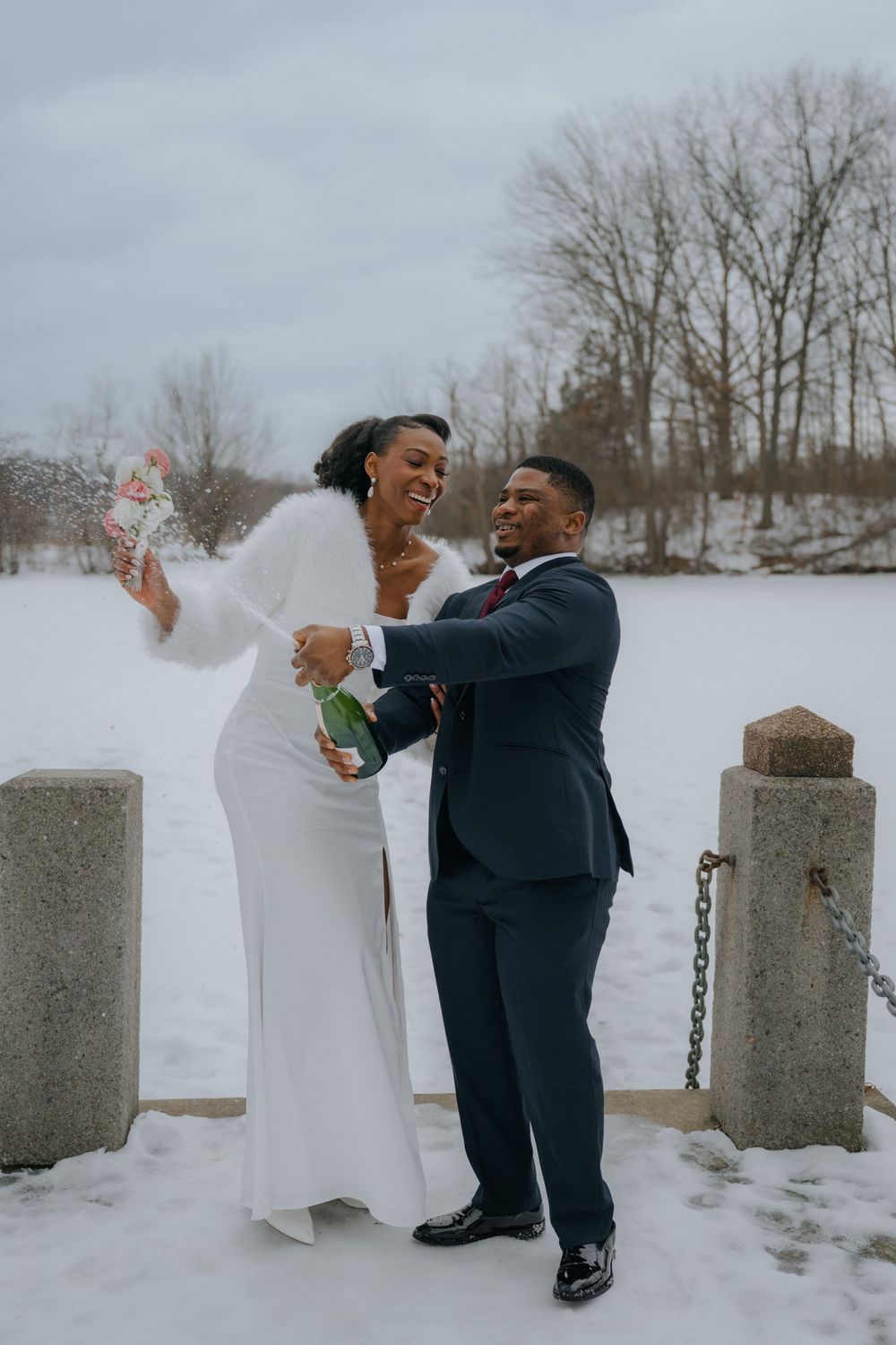 A couple in formal attire dances playfully in the snow near stone pillars during a winter photo session.