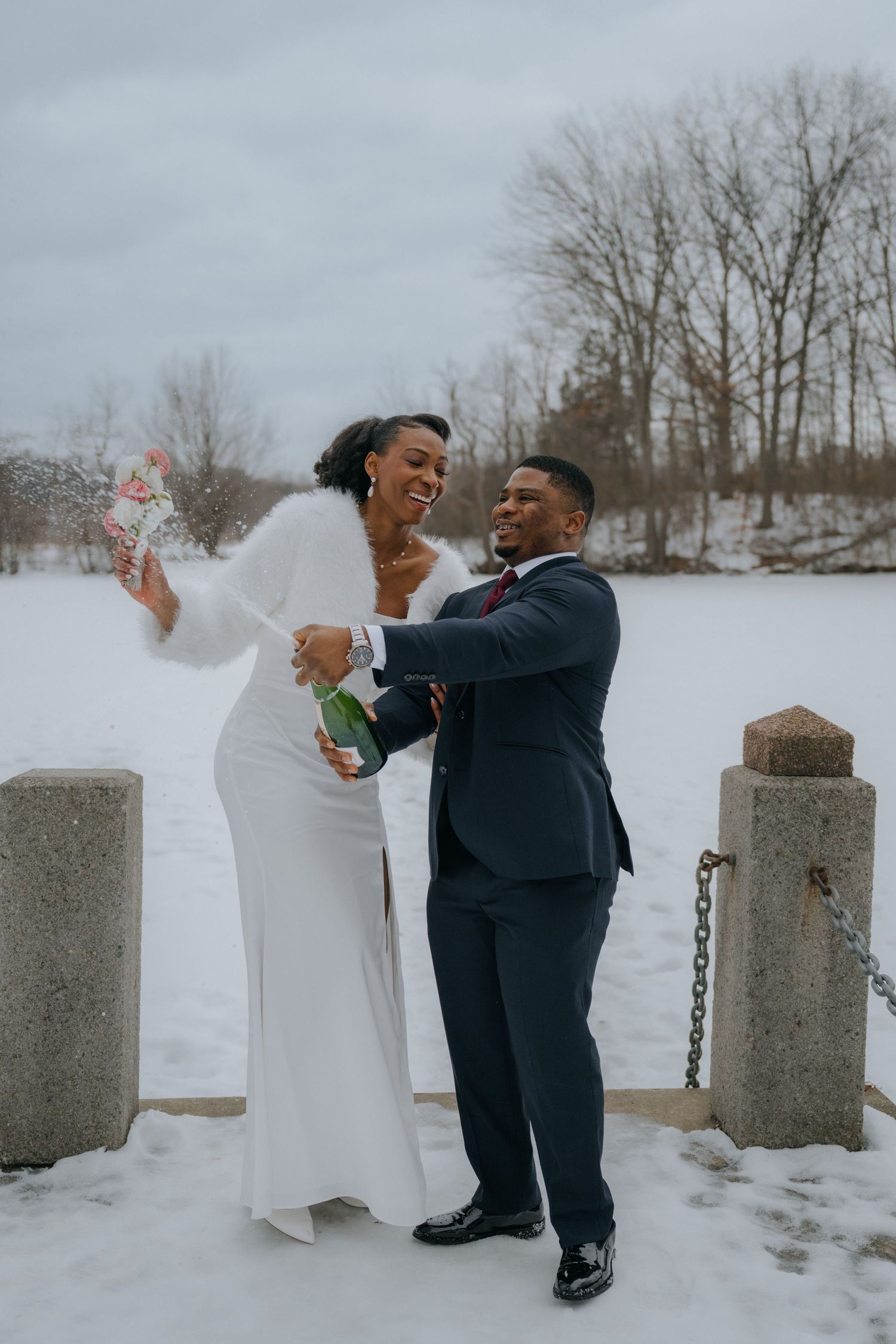 A couple in formal attire dances playfully in the snow near stone pillars during a winter photo session.