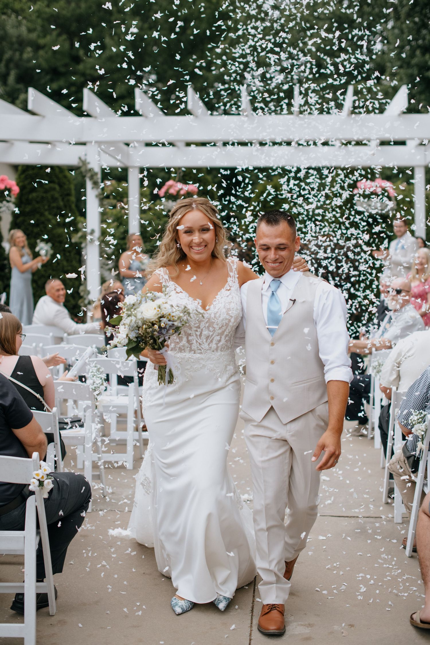 Newlyweds walking down the aisle under a white pergola as confetti rains down during outdoor ceremony.