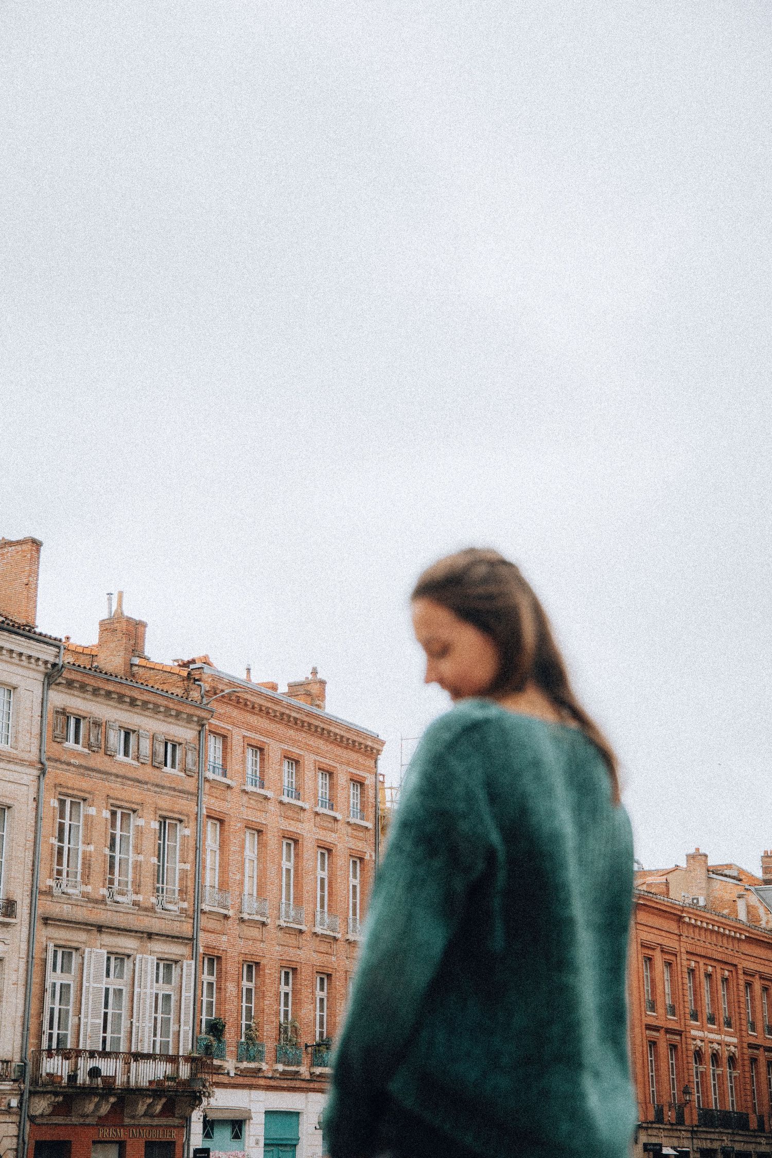 A young woman in a teal knit sweater stands in front of classic European buildings on an overcast day.