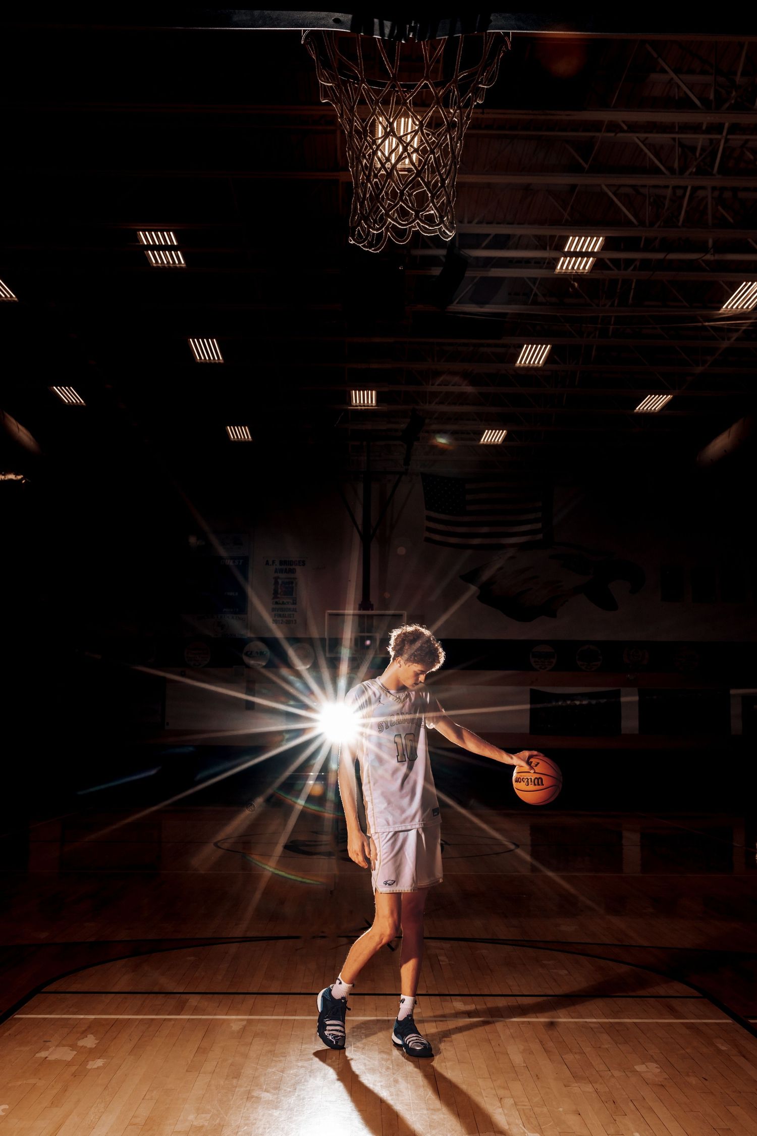 Basketball player dribbling on wooden court with dramatic overhead lighting creating dynamic shadows and lens flare effects.