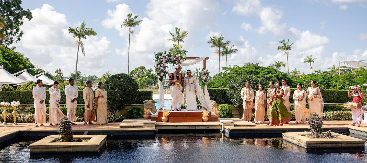 Outdoor Indian wedding ceremony beside a pool with wedding party in traditional attire under a floral mandap.