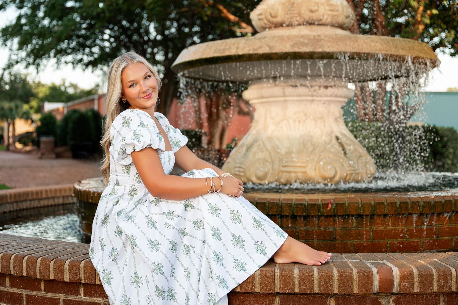 Woman in white floral dress poses by brick fountain in outdoor garden setting.