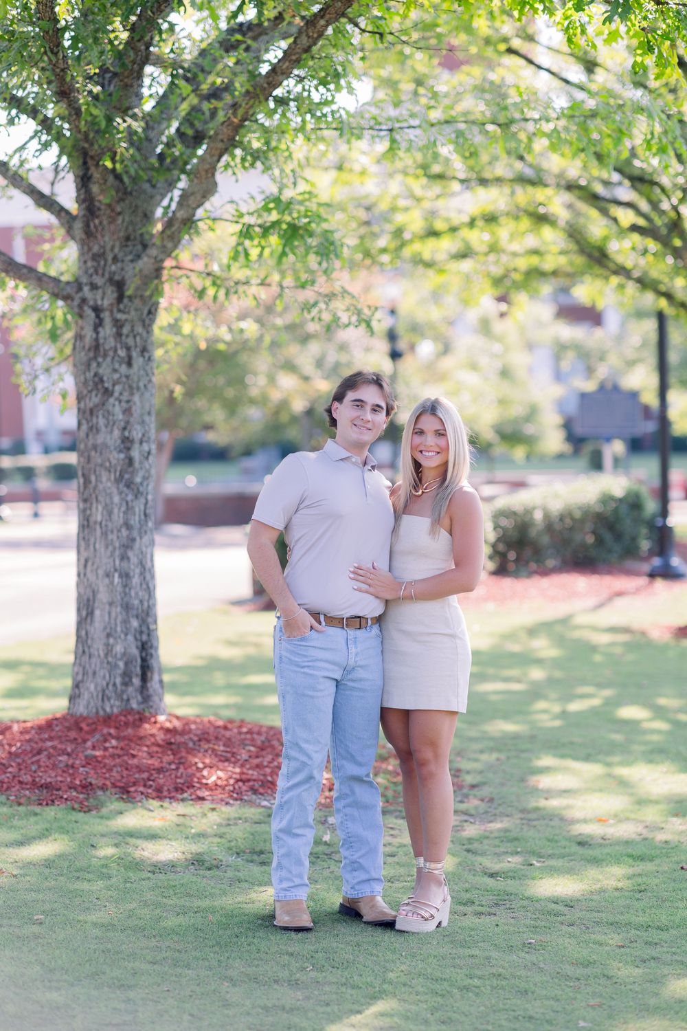 A couple poses together on a sunny day under trees with red mulch and green grass in a park-like setting.