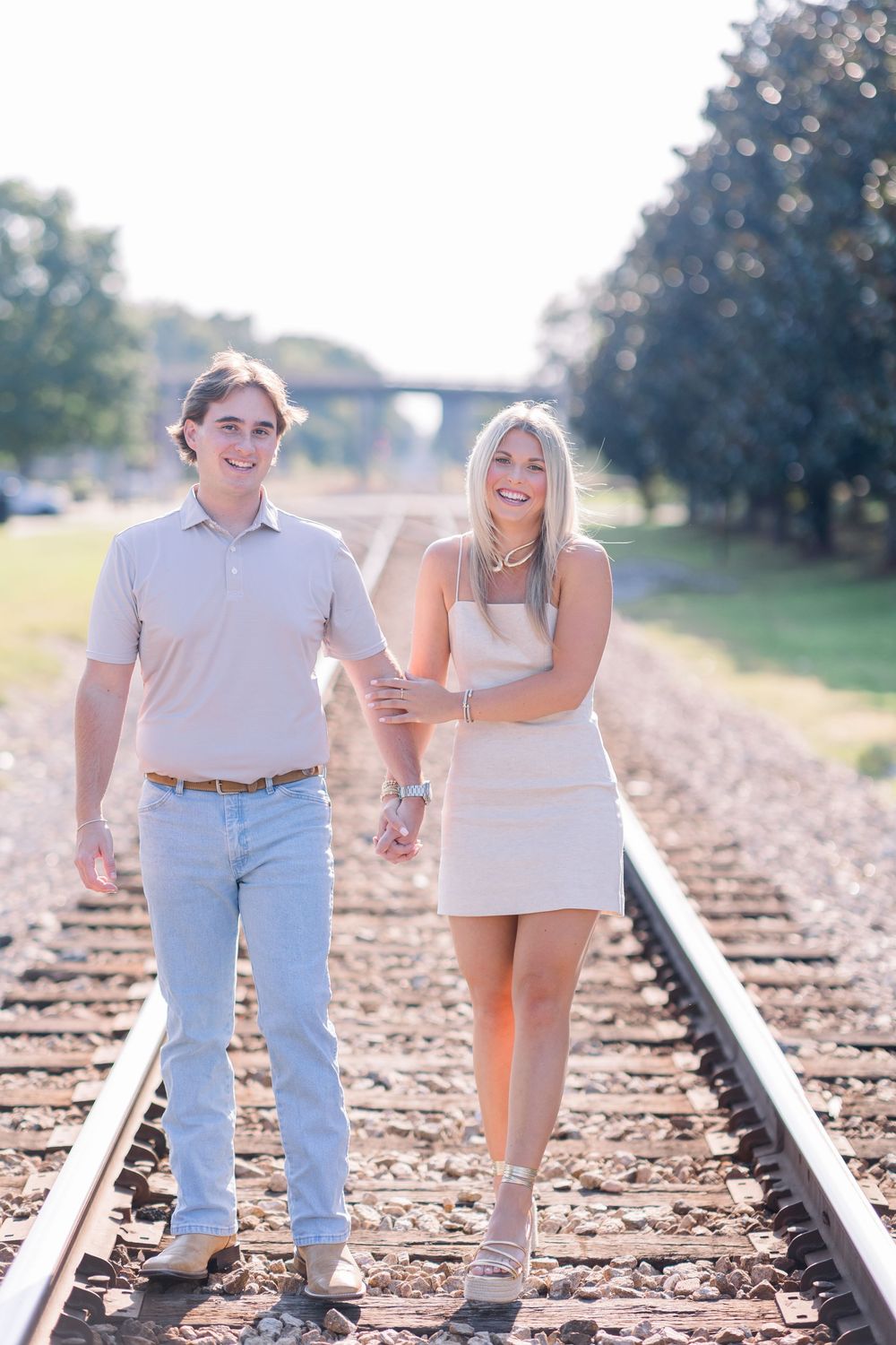 A couple walks hand in hand along railroad tracks on a sunny day, wearing light colored casual outfits.