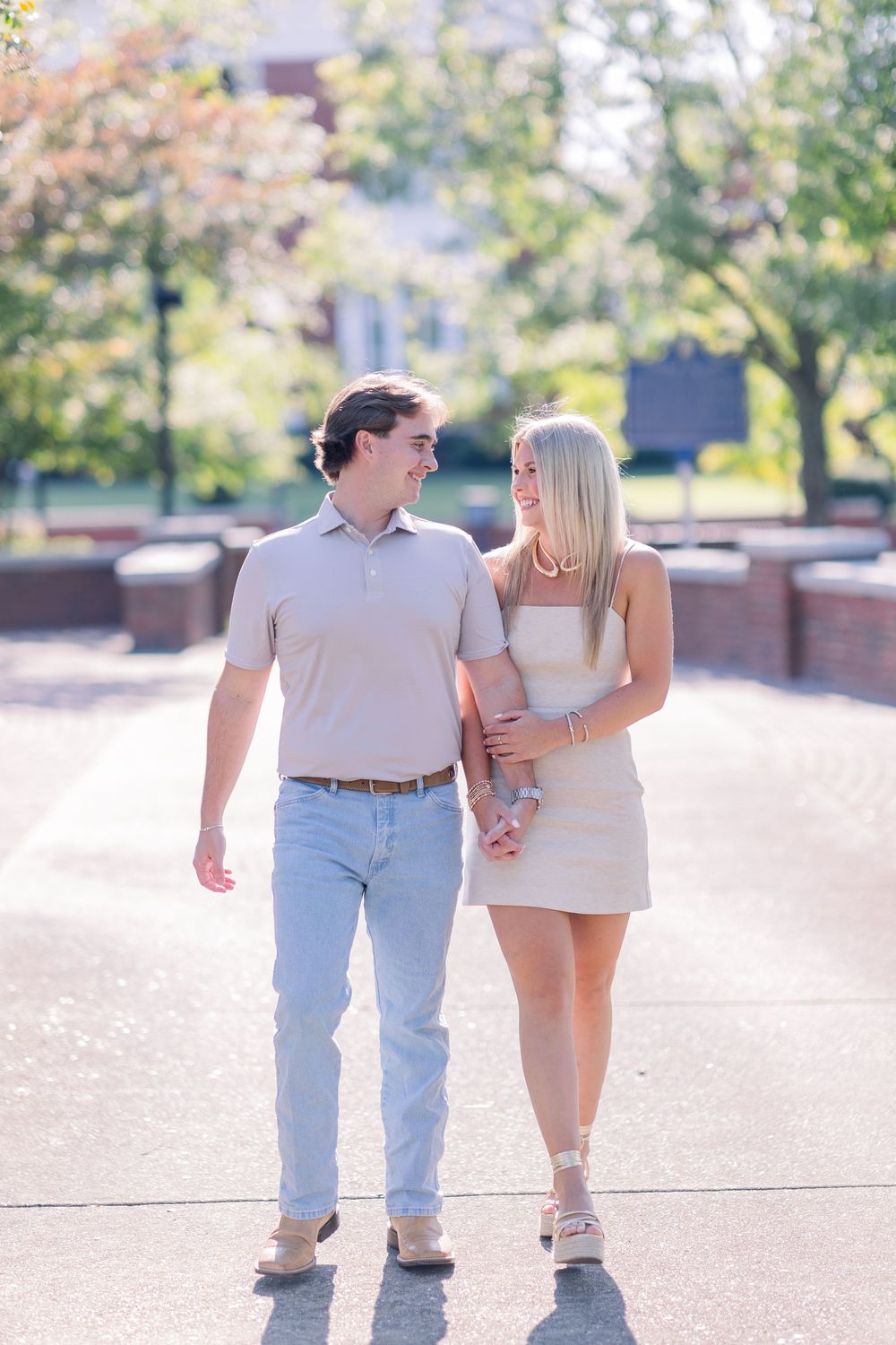 A couple in casual summer attire walking together on a sunny paved path with trees and bokeh lighting in the background.