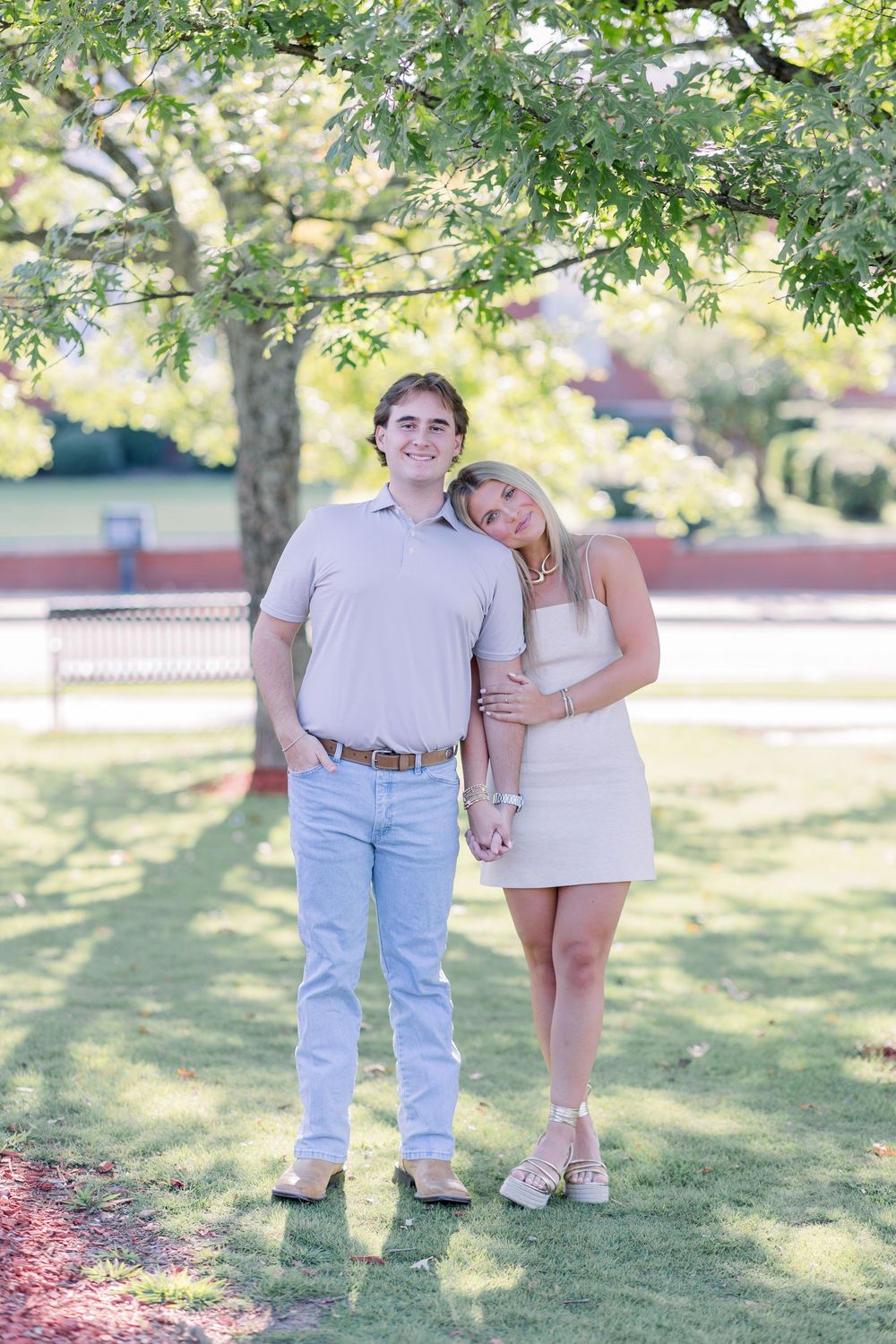 A couple poses playfully in a sunny park setting with green trees in the background.