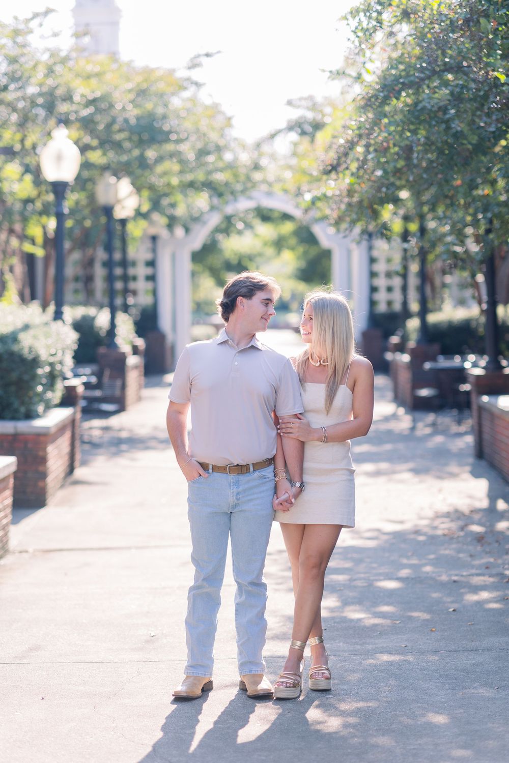 A couple walking hand in hand along a tree-lined brick pathway in casual summer attire.