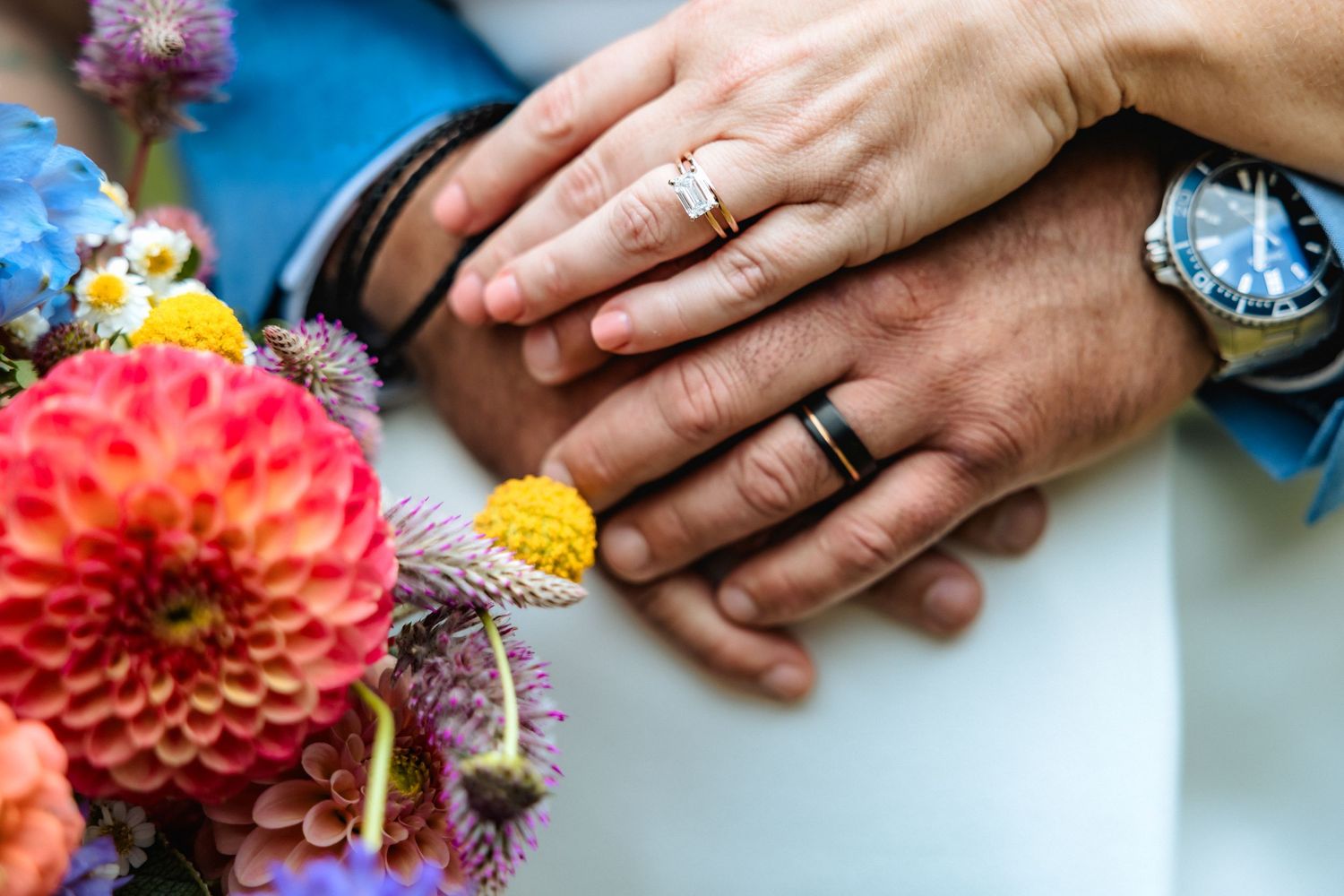 Hands with wedding rings rest near vibrant dahlias and colorful flowers arranged on a light surface.