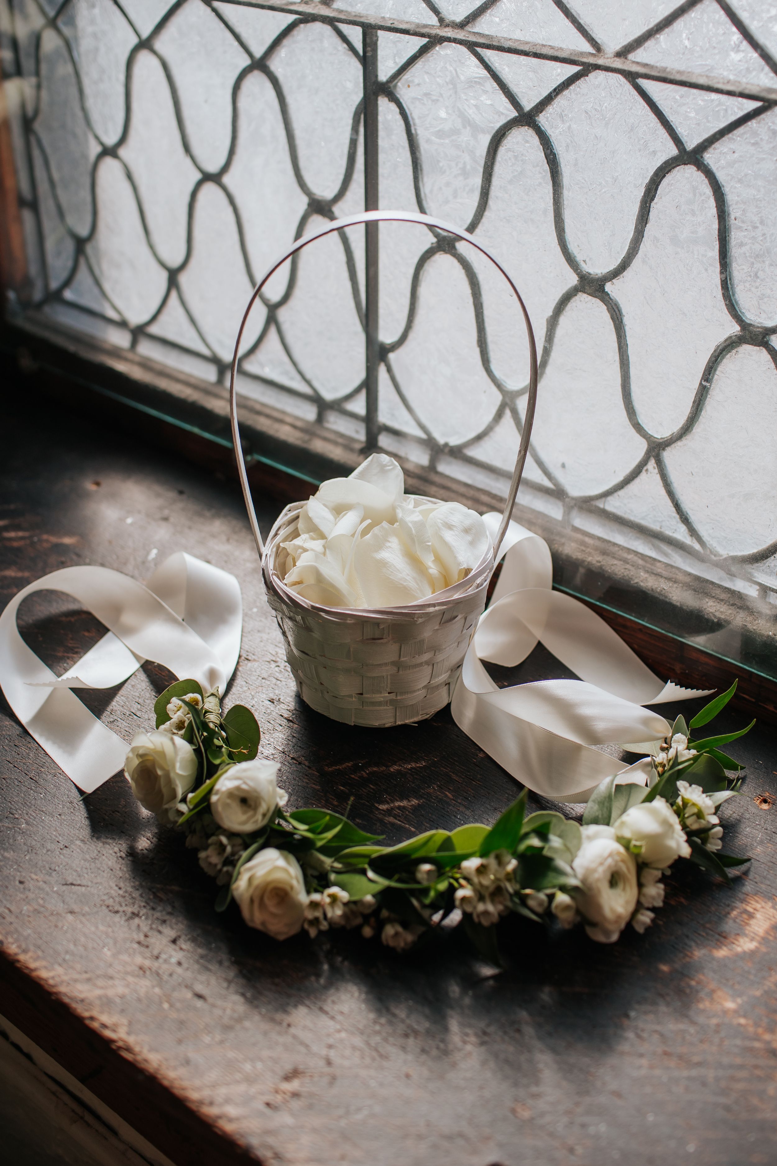 White basket filled with delicate fabric alongside elegant white roses and ribbons on a rustic wooden surface by a window.