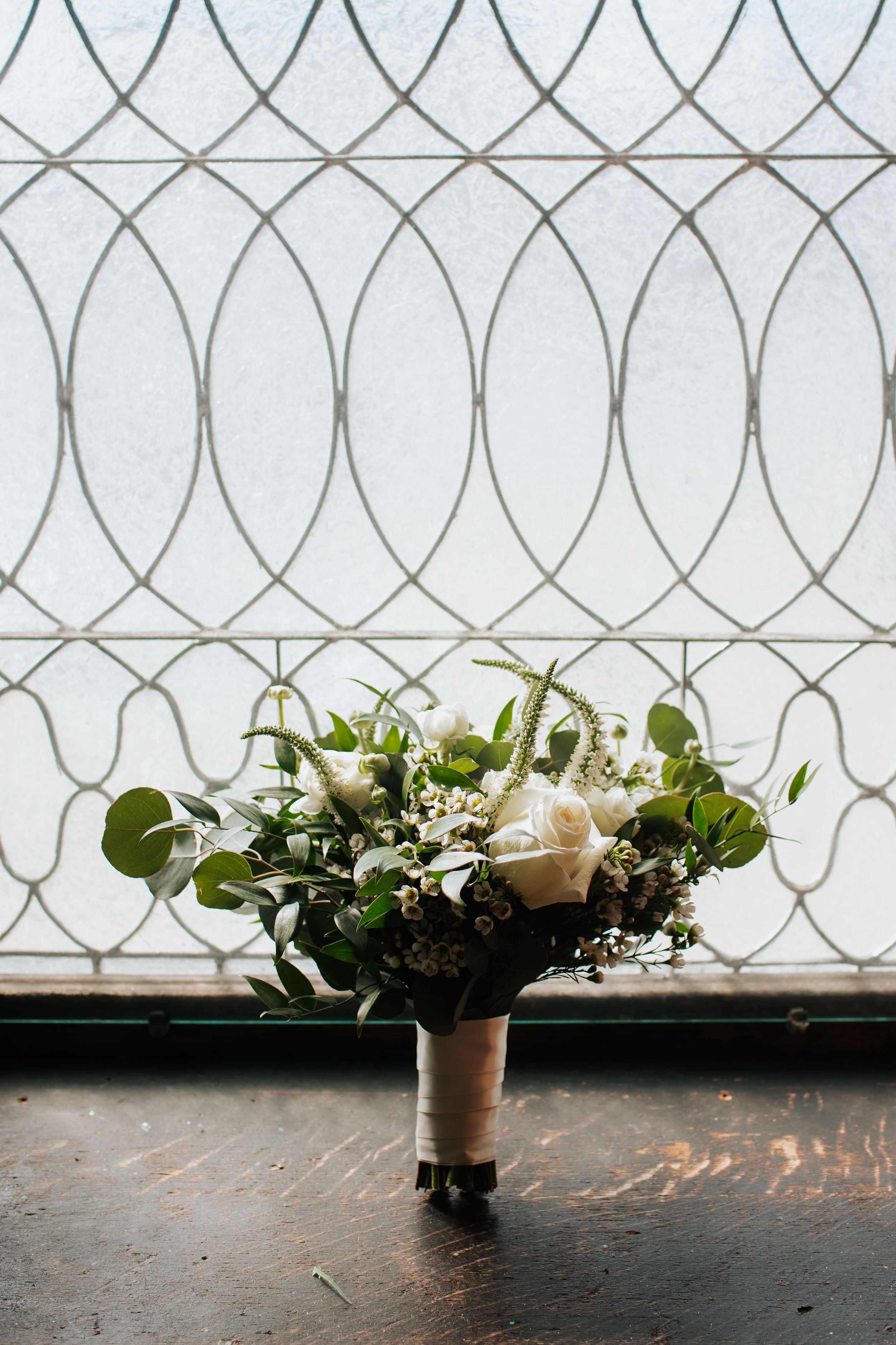 A rustic wedding bouquet with white roses and greenery sits against a decorative window with geometric pattern background.