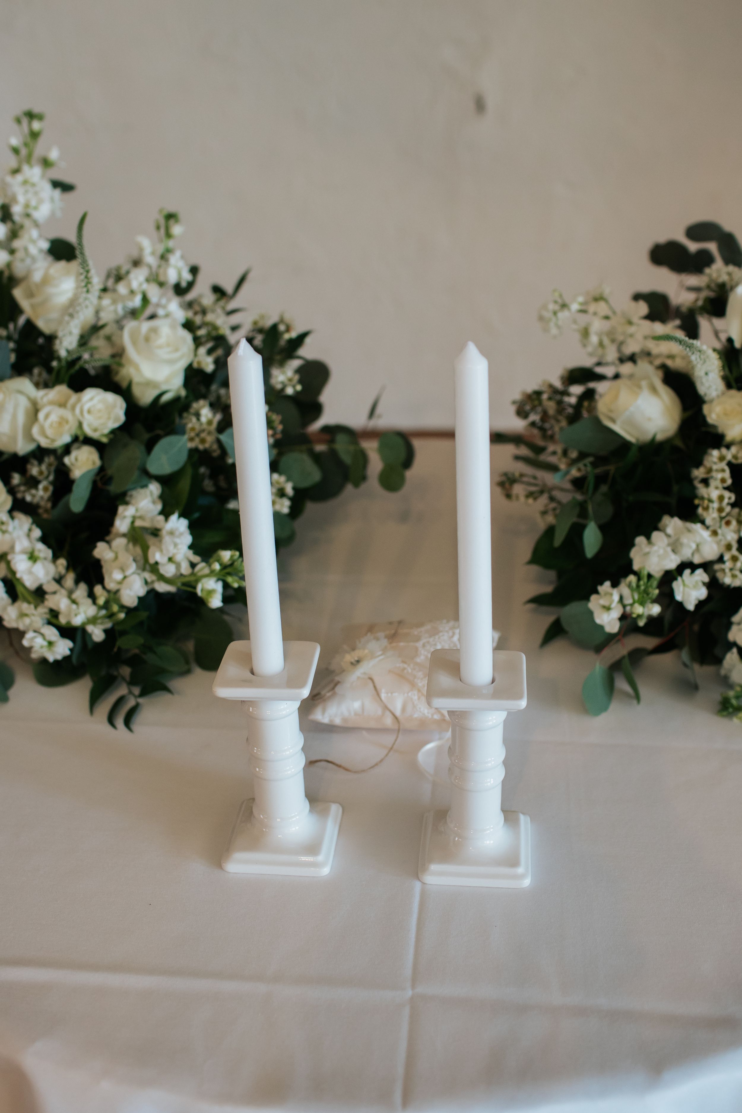 White taper candles in ceramic holders sit beside elegant floral arrangements with white blooms and greenery on a table.