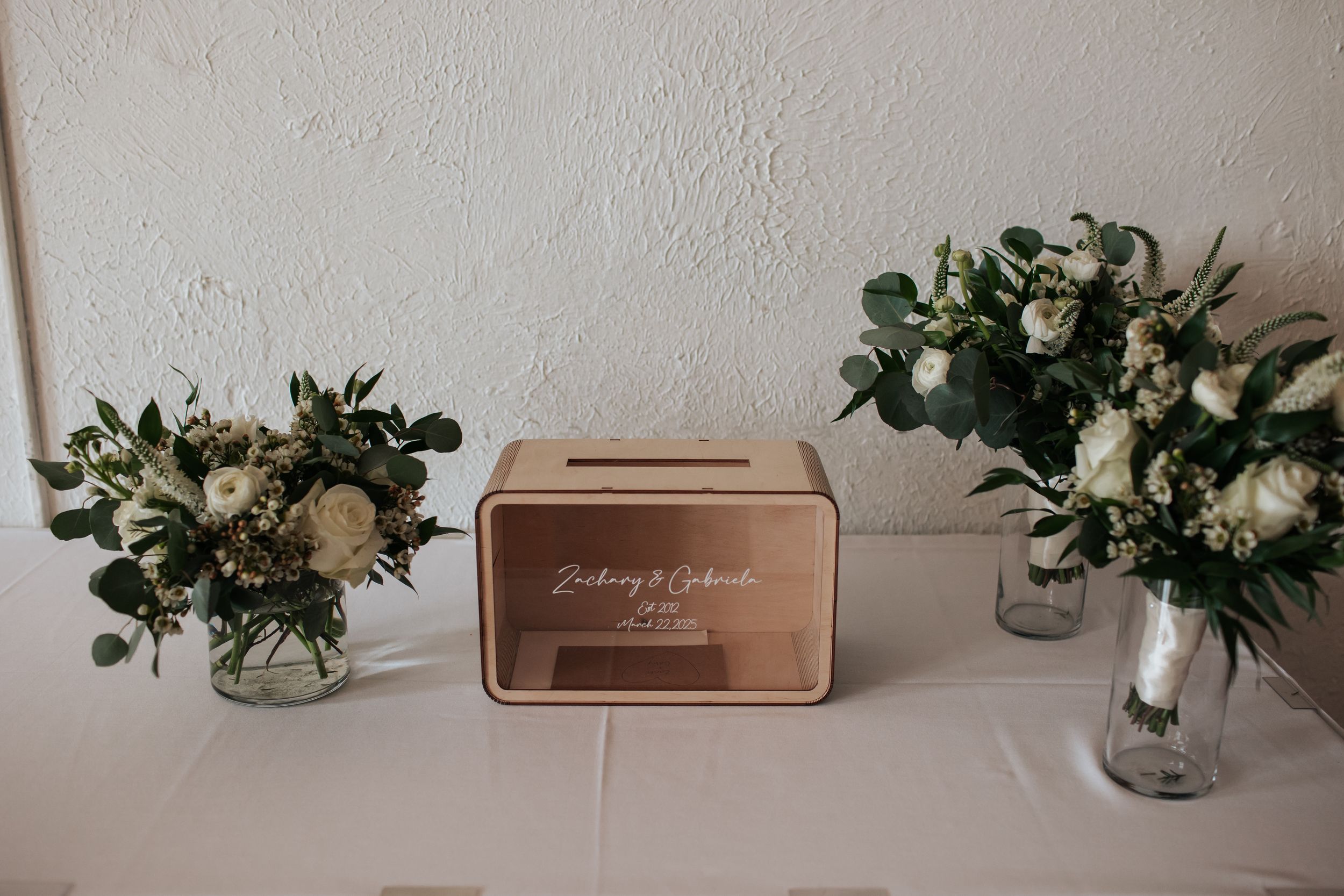 A row of elegant floral centerpieces with white flowers and greenery in glass vases line a white surface.