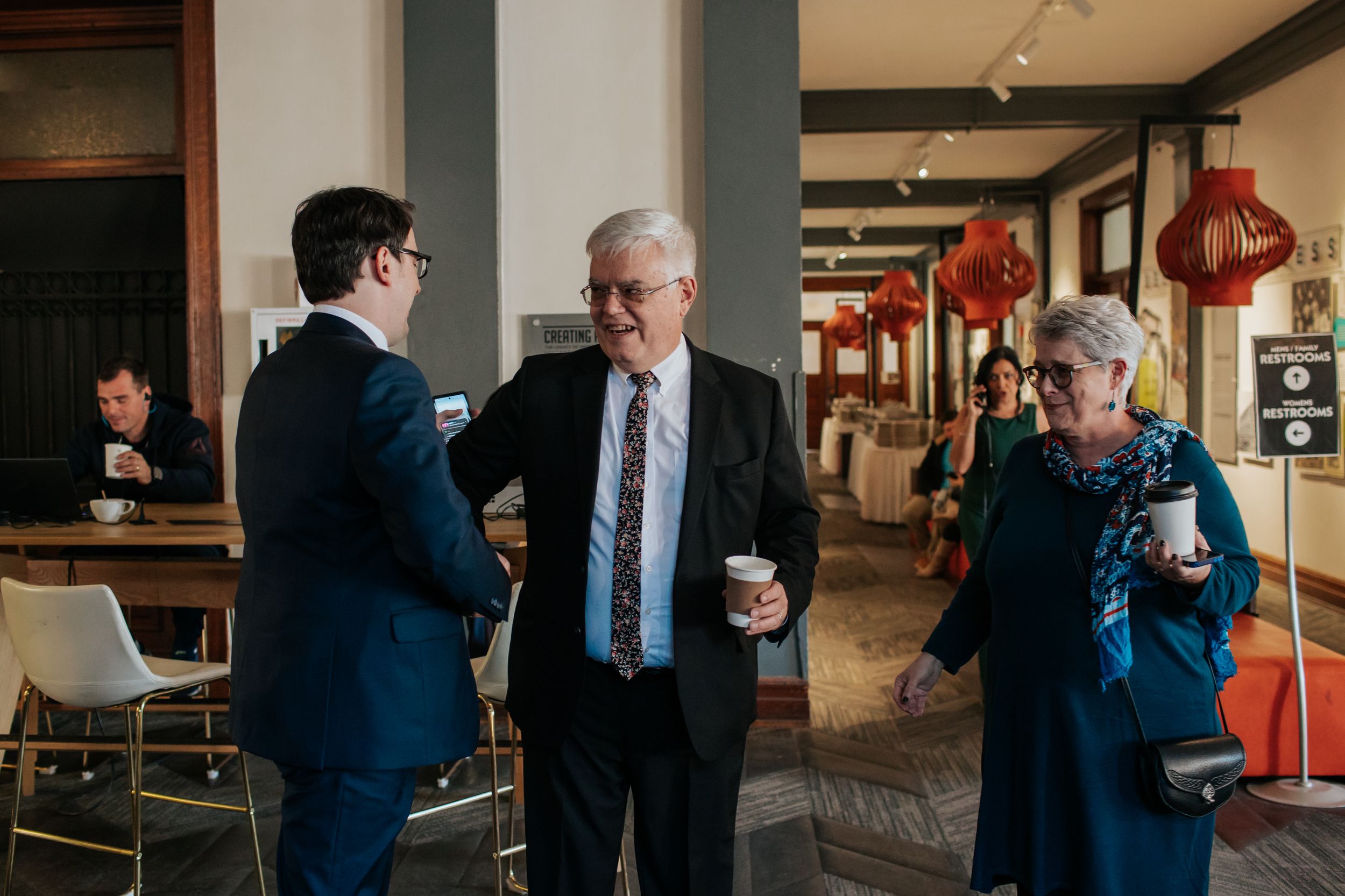 Two people stand in conversation while one holds a coffee cup in a modern office hallway setting.